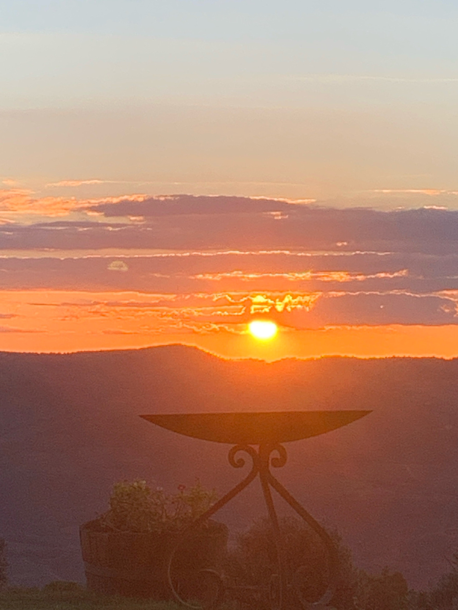 The image depicts a sunset over the landscape of Castiglione d'Orcia, Italy. The sun, a bright orb, is partially obscured by clouds, casting a warm orange glow across the sky. The foreground features a decorative metal stand with a wide, shallow basin, possibly for a fire pit. To the left of the stand, a large, dark pot contains green plants and a few small red flowers. The background consists of rolling hills and mountains, silhouetted against the vibrant sky. The overall scene is peaceful and serene, capturing the natural beauty of the Tuscan countryside at dusk.