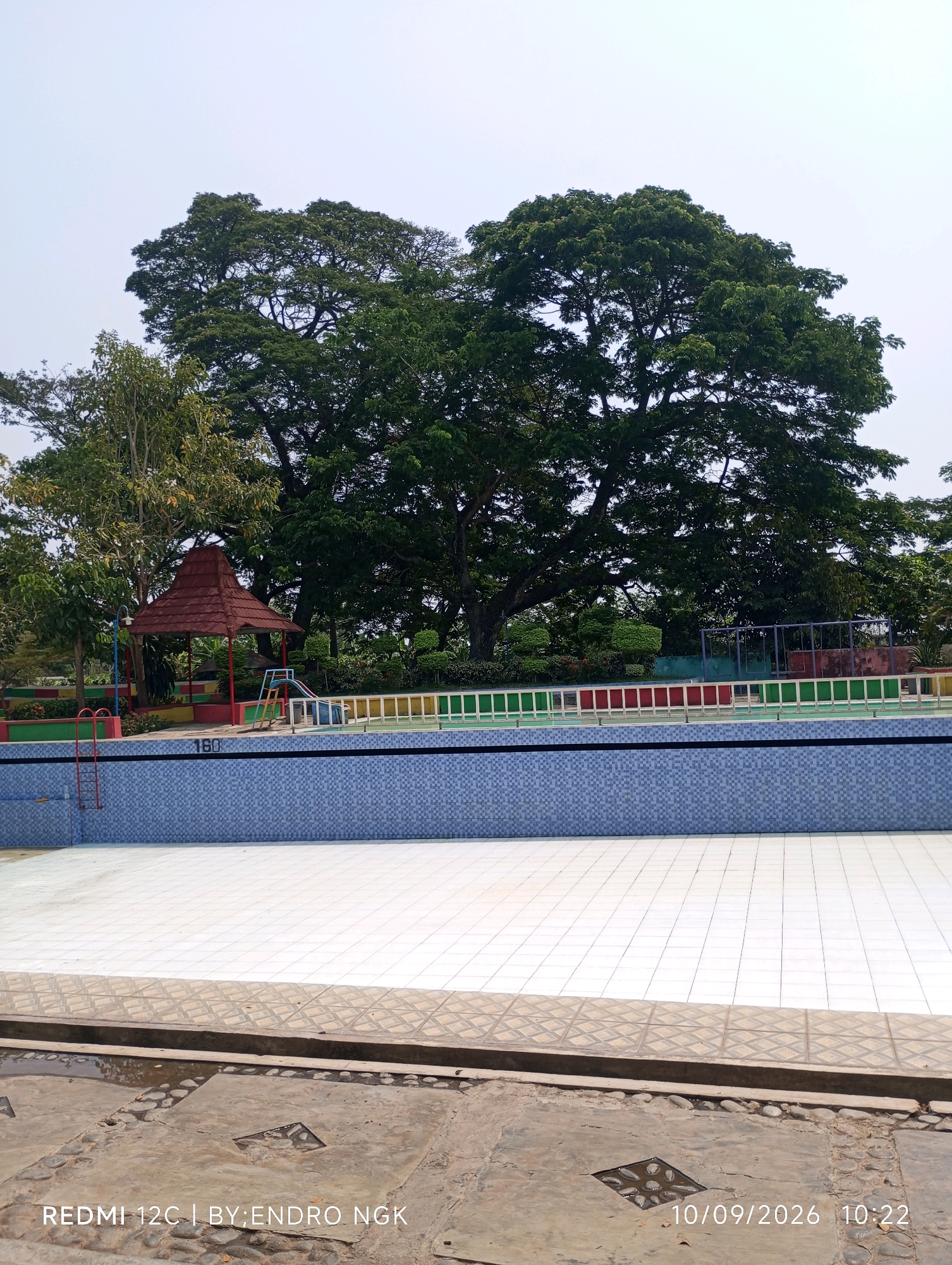 An outdoor swimming pool in Sombron, Indonesia, features a blue tiled deep end and a white tiled shallow end. A red ladder is visible on the left side, with a sign displaying "160" on the pool's edge. In the background, a red-roofed gazebo stands amongst lush green trees. A colorful fence separates the pool area from the surrounding greenery. The sky is overcast. The bottom of the image contains text identifying the device used, the photographer, and the date and time the photo was taken.