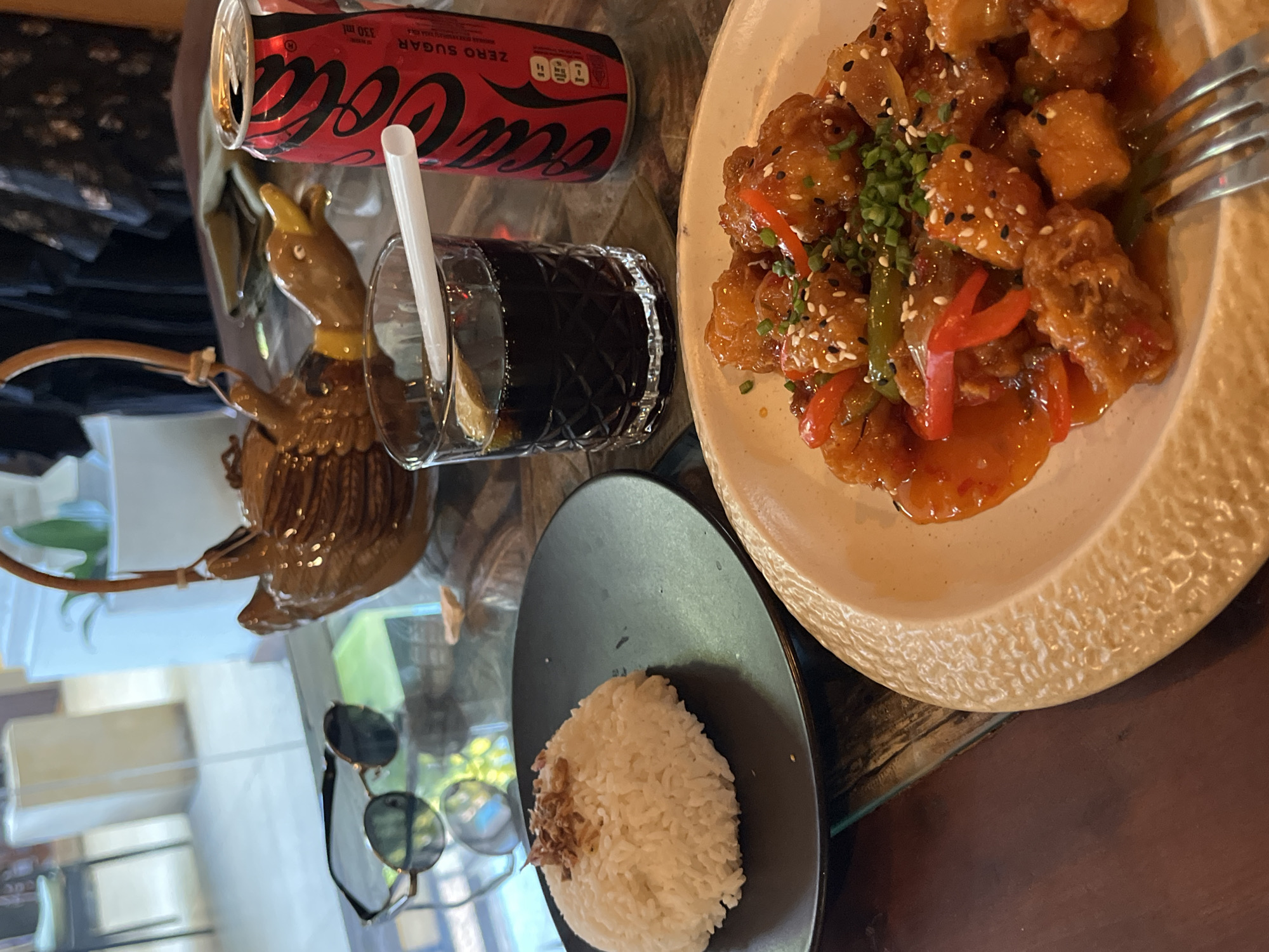 The image captures a dining table scene. There is a plate of sweet and sour chicken, sprinkled with sesame seeds and garnished with chopped green onions, is placed on the table along with a side plate of white rice with a small amount of topping. A can of Coca-Cola Zero Sugar and a glass of dark beverage with a straw are also present. An ornamental teapot-shaped object sits nearby. In the lower left corner, a pair of sunglasses rests on the table. The scene appears to be inside a restaurant or cafe, possibly in Kuta Selatan, Indonesia. The lighting suggests a daytime setting. No people are visible.