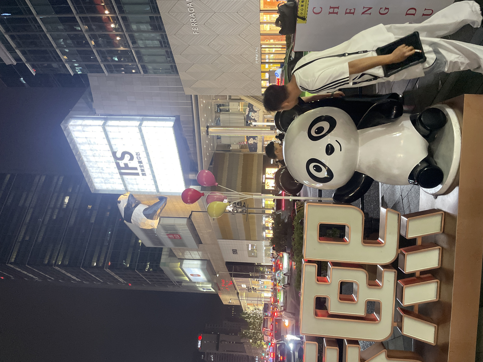 At night in Jinjiang District, China, a man in a white Adidas tracksuit, with a black bag, uses a tablet. The man is next to a large panda statue wearing a pair of black shorts. Next to the panda is a large, lighted, red-outlined sign with white characters. In the background are buildings, including the IFS building. Red, pink, and yellow balloons float above the panda statue. Cars and pedestrians are visible on the street. The Ferragamo store is also visible.