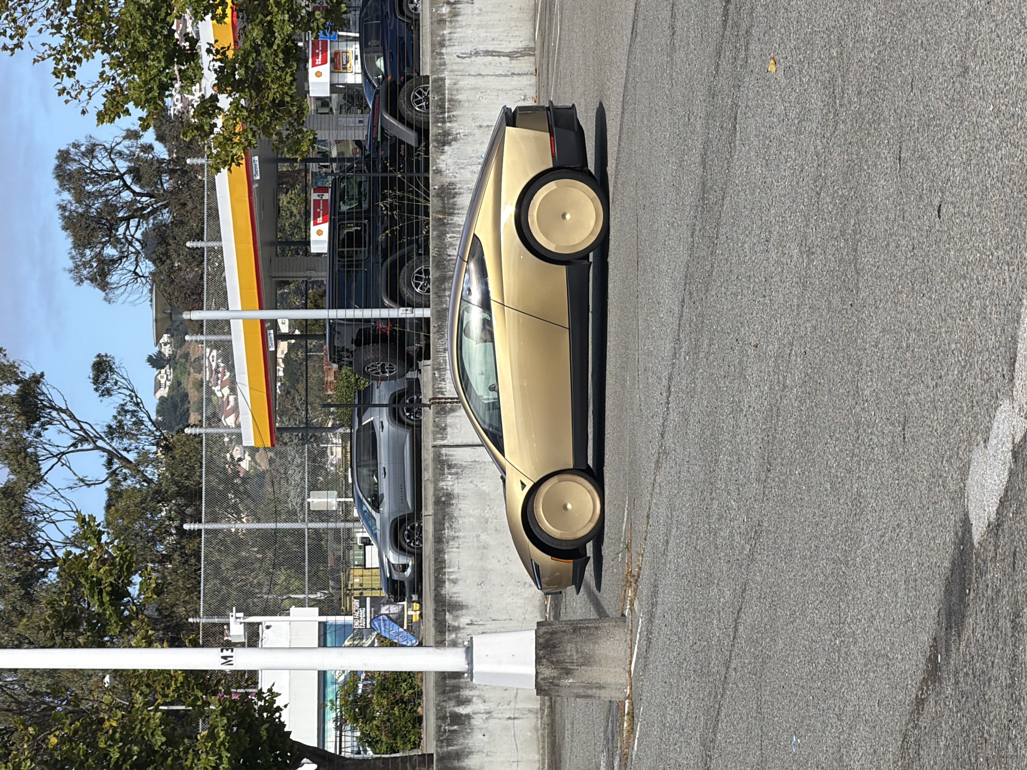 The media file depicts a side view of a futuristic, light gold car parked on an asphalt surface. The car has a sleek design with a unique, flat-topped wheel on the visible rear wheel. Its windows are tinted, and the overall aesthetic suggests a modern electric vehicle. The car is positioned in what appears to be a parking lot or roadside area.
Behind the car, a concrete wall runs horizontally, and beyond that, a chain-link fence encloses a view of several other vehicles parked in a row. These vehicles include a dark blue SUV and a silver SUV, with some hinting at a Jeep brand. A Shell gas station canopy with its signature red and yellow branding is visible in the background, suggesting the location is near a gas station. Trees with green foliage are present on the left side of the image, and a few buildings are visible on a distant hill.
The lighting indicates it is daytime, with shadows cast by the car and other objects, suggesting either late morning or afternoon. The sky is clear and blue. The presence of the gas station and the variety of vehicles imply a public or commercial setting, possibly a retail area or a service station.
There is some visible text in the image, though it's partially obscured and rotated. On a white pole in the foreground, text reads "ME" vertically, likely part of a larger word or sign. In the background, near the fence, a sign reads "END FACTORY PARKING". The overall scene conveys a sense of everyday activity in a suburban or commercial environment under clear weather conditions.