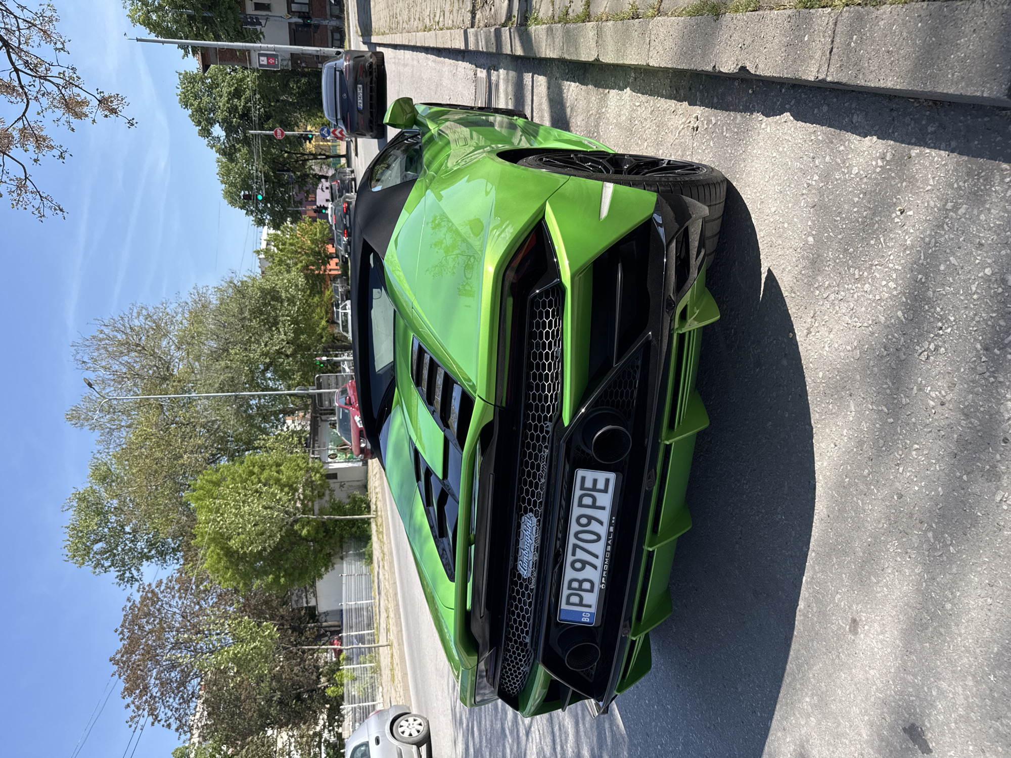 The primary subject of the image is a bright green Lamborghini Huracán convertible, viewed from its rear-right quarter, on an urban street in Plovdiv, Bulgaria. The supercar is striking with its vivid lime green metallic paint, aggressive aerodynamic lines, large rear diffuser fins, and prominent twin exhaust pipes. It features black honeycomb grilles and black multi-spoke wheels.
The car appears to be stationary, either parked along the curb or stopped in traffic. In the background, other vehicles are visible, including a dark sedan with a Bulgarian license plate (PA 3475 HE) and a silver car partially visible on the left. The scene is set on a paved road with a concrete curb and sidewalk running parallel to the car.
Notable details include the clear blue sky, indicating a sunny day. Strong shadows are cast by the Lamborghini and nearby trees, suggesting it is mid-morning or afternoon. Lush green trees line the street and are abundant in the background, along with some distant buildings. Traffic signals are visible further down the road, with one showing a green light. A red octagonal "STOP" sign and a blue circular traffic sign with a white arrow (likely indicating a mandatory turn or direction) are mounted on poles.
Visible text includes the Bulgarian license plate "PB 9709 PE" on the rear of the Lamborghini, with the country code "BG" on the blue EU strip. The text "PROMOSALE.BG" is visible on the license plate frame. The "Lamborghini" script emblem is centered above the license plate. The word "STOP" is clearly legible on the red octagonal sign.