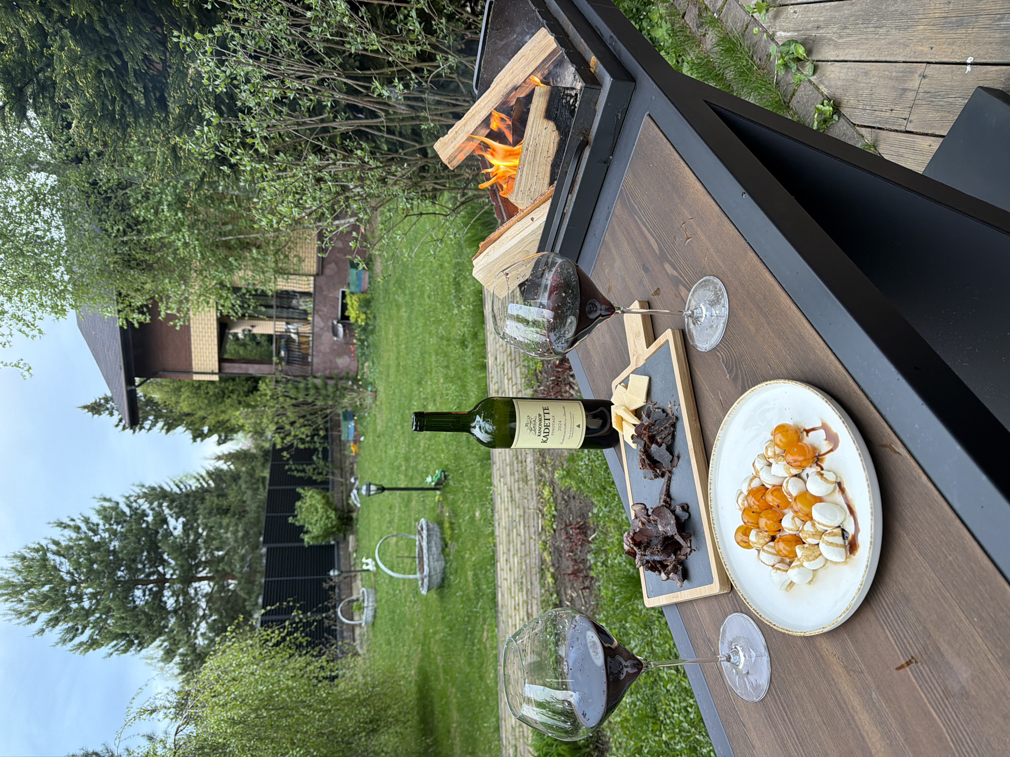 The image shows a picnic or gathering in an outdoor setting, likely a backyard or patio. A fire is burning in a metal fire pit that doubles as a table. On the table, there are two wine glasses filled with red wine, a bottle of wine, a cutting board with cheese and dried meat, and a plate of small white and orange balls drizzled with a dark sauce, probably mozzarella and tomatoes. The environment includes green grass, trees, a brick walkway, a black fence, and a house in the background. The sky is overcast. The location is not definitively identifiable as Zimyonki, Russia, based on the provided imagery. There are no people visible in the shot.