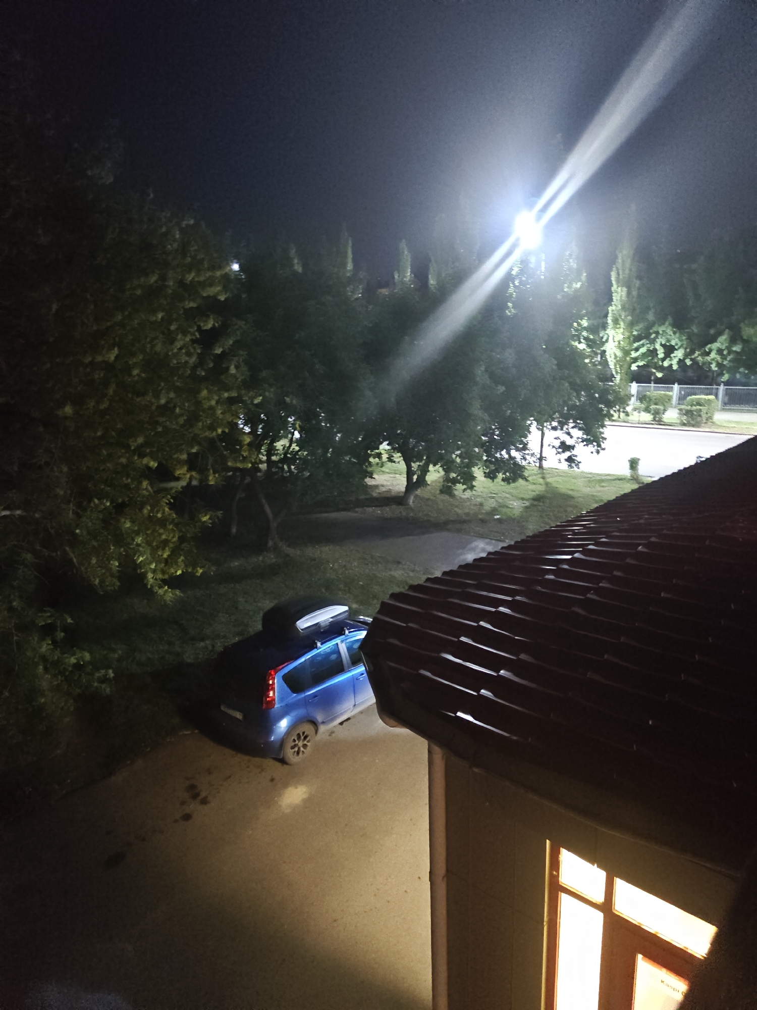 An outdoor, overhead, night-time shot shows a blue hatchback parked on an asphalt surface. The car has a black roof box. To the left of the car is a grassy area and a large, leafy tree. Behind the tree is another tree and a bright, overhead street light. In the background is a fence and more trees. In the foreground, is a red tiled roof. The scene is likely in a residential area in Petropavl, Kazakhstan.