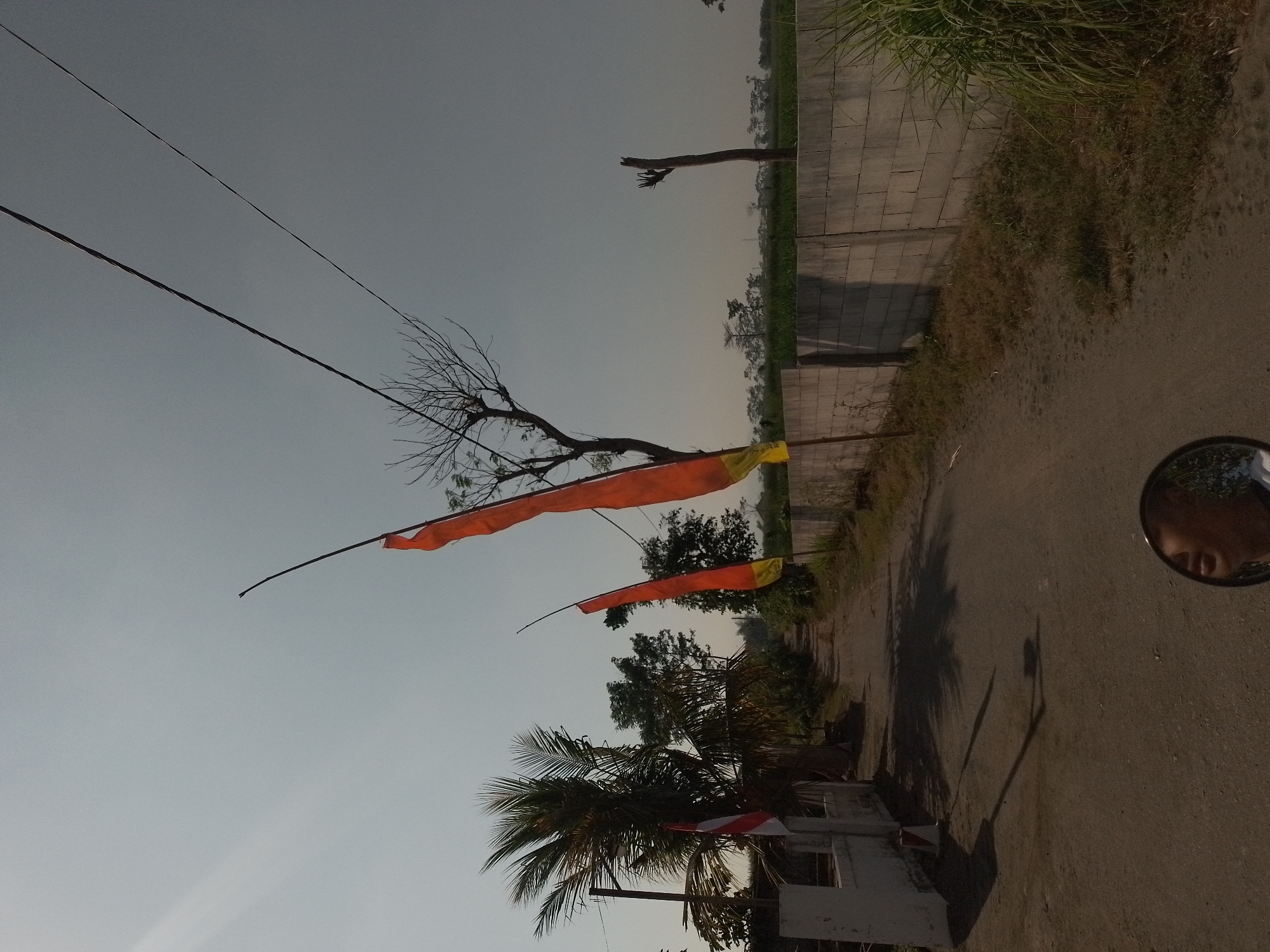 The image depicts a rural scene on a sunny day, likely in or near Ngancar, Indonesia, given the presence of national flags. The viewpoint is from a vehicle, as evidenced by the reflection of a person's face and the vehicle's mirror in the lower part of the frame. A dirt road stretches into the distance, flanked by a low, concrete block wall on the right and a white-painted structure on the left. Several tall, orange and yellow flags are visible, attached to poles, swaying in the breeze. One of these flags, on the left side of the road near the white structure, appears to be a red and white Indonesian flag. The environment includes scattered trees, one with bare branches, and a flat expanse of green vegetation in the background. Overhead, power lines crisscross the sky, which is a pale blue with some hazy sunlight. The overall impression is of a quiet, rural roadside.