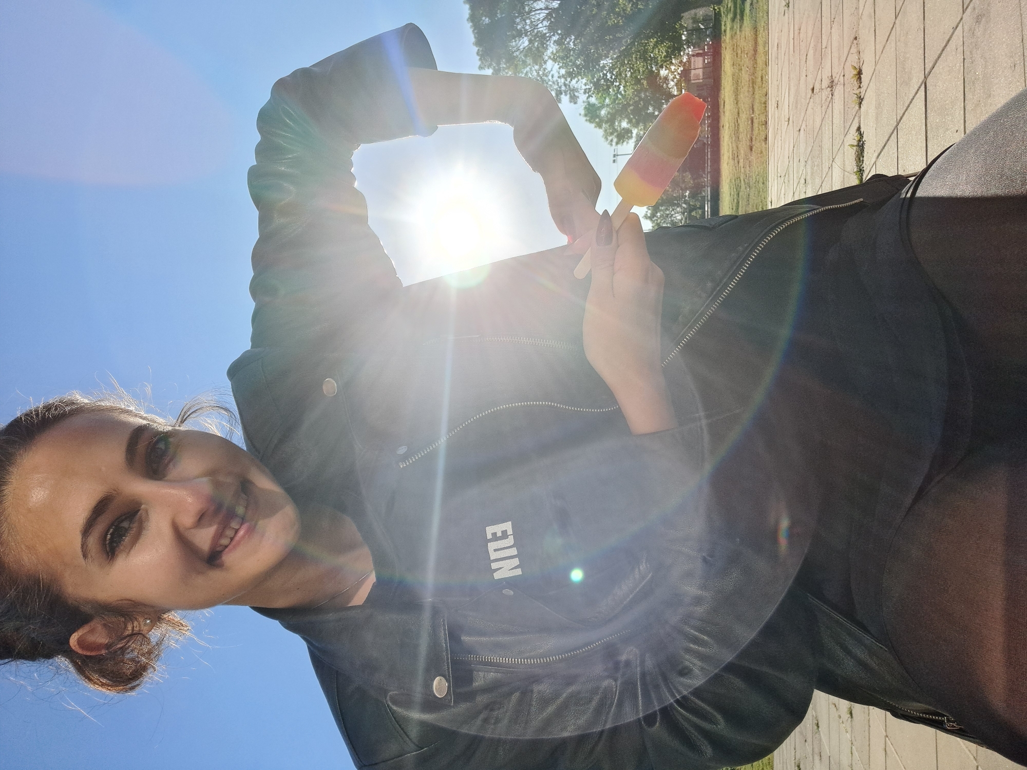 A woman holds a multi-colored popsicle in her right hand while framing the sun with her left hand. She is smiling at the camera. The word "FUN" is visible on her black leather jacket. The location appears to be outdoors on a paved surface in a city park, on a sunny day.