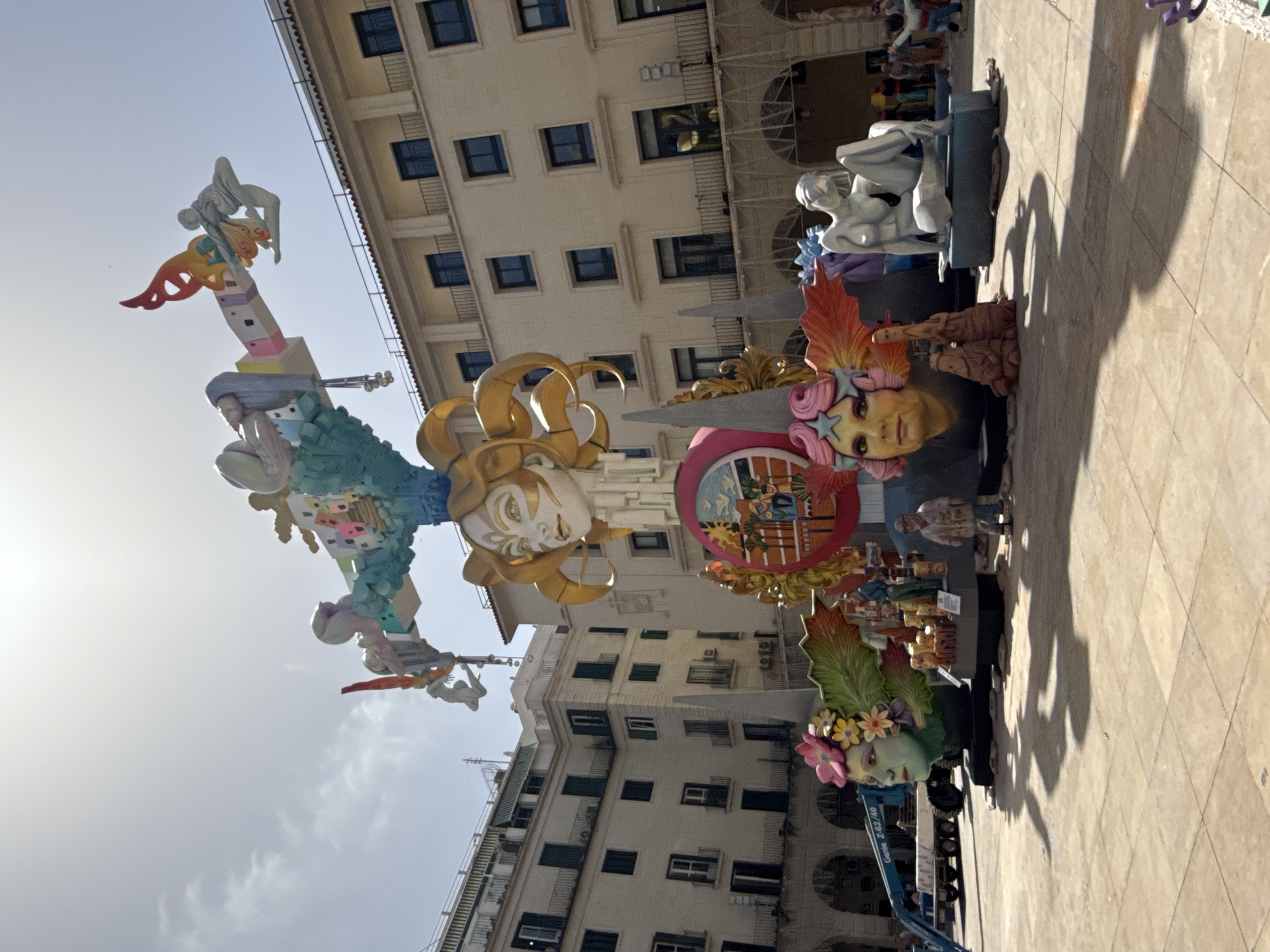 An outdoor, eye-level, wide-angle photograph captures a street scene in front of cream-colored, multi-story buildings with dark windows and balconies. The sky is bright and partly cloudy. Dominating the center of the image is a large, elaborate sculpture, possibly part of a festival or parade. The sculpture features several large, stylized faces and figures, with vibrant colors and intricate details. In the foreground, a large, green face adorned with flowers is visible, with a smaller, pink-haired face to its right. A light blue Genie lift is parked to the left of the green face. In the mid-ground, the sculpture continues upwards with more faces and abstract elements, including a golden-horned face at the top. To the right of the sculpture, a white, seated nude figure is positioned on a pedestal. Several smaller figures and structures are incorporated into the sculpture's base. People are visible in the background, some standing and observing the sculpture, and others appearing to be involved in the setup or dismantling of the display. The ground is paved with large, square tiles. Shadows are cast across the paved area, indicating a sunny day.