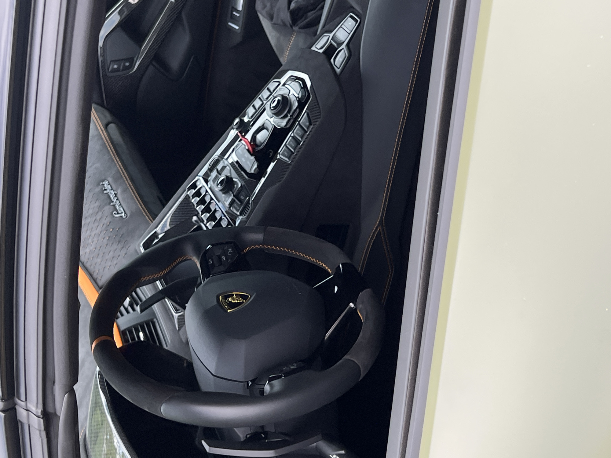 The image shows the interior of a car, specifically the dashboard and steering wheel. The steering wheel is black with a yellow Lamborghini emblem. The dashboard is a glossy material and includes several buttons and knobs. The car's interior appears to be in pristine condition. The location is not discernible.