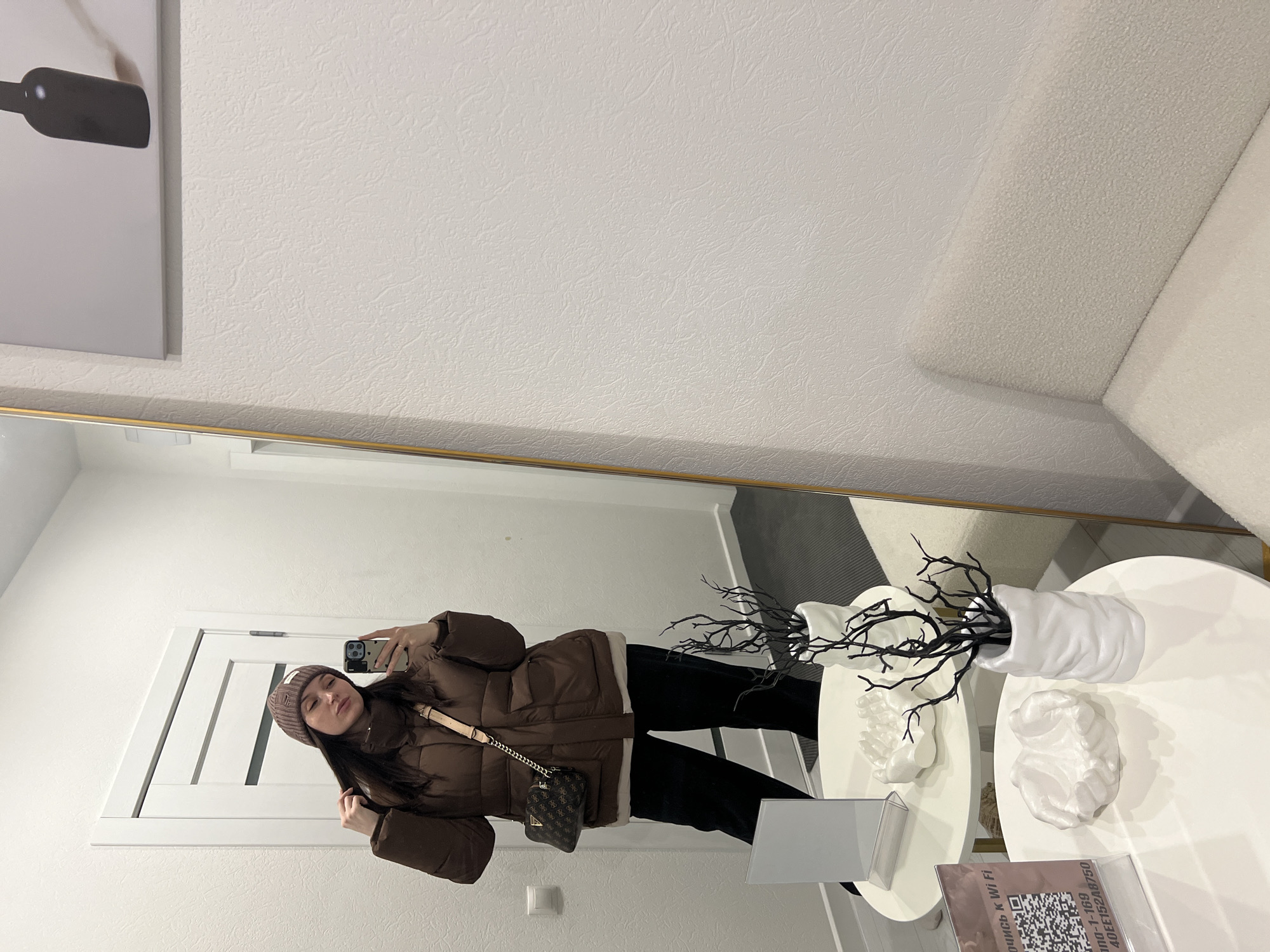 A young woman wearing a brown beanie, brown puffer jacket, and black pants is taking a selfie in a mirror. She holds a smartphone in her right hand, capturing her reflection. A small, brown handbag hangs from her shoulder.
The reflection shows an interior space with white walls and a white door. A piece of art depicting a bottle hangs on the wall behind her. To the right, a white sofa is partially visible. A small, white, round table stands in front of the mirror with decorative items, including a white vase with black twigs and two white hand sculptures. A sign with a QR code is on the table, indicating possible Wi-Fi information.
Given the signage, the scene is likely in a commercial space in Tyumen, Russia.