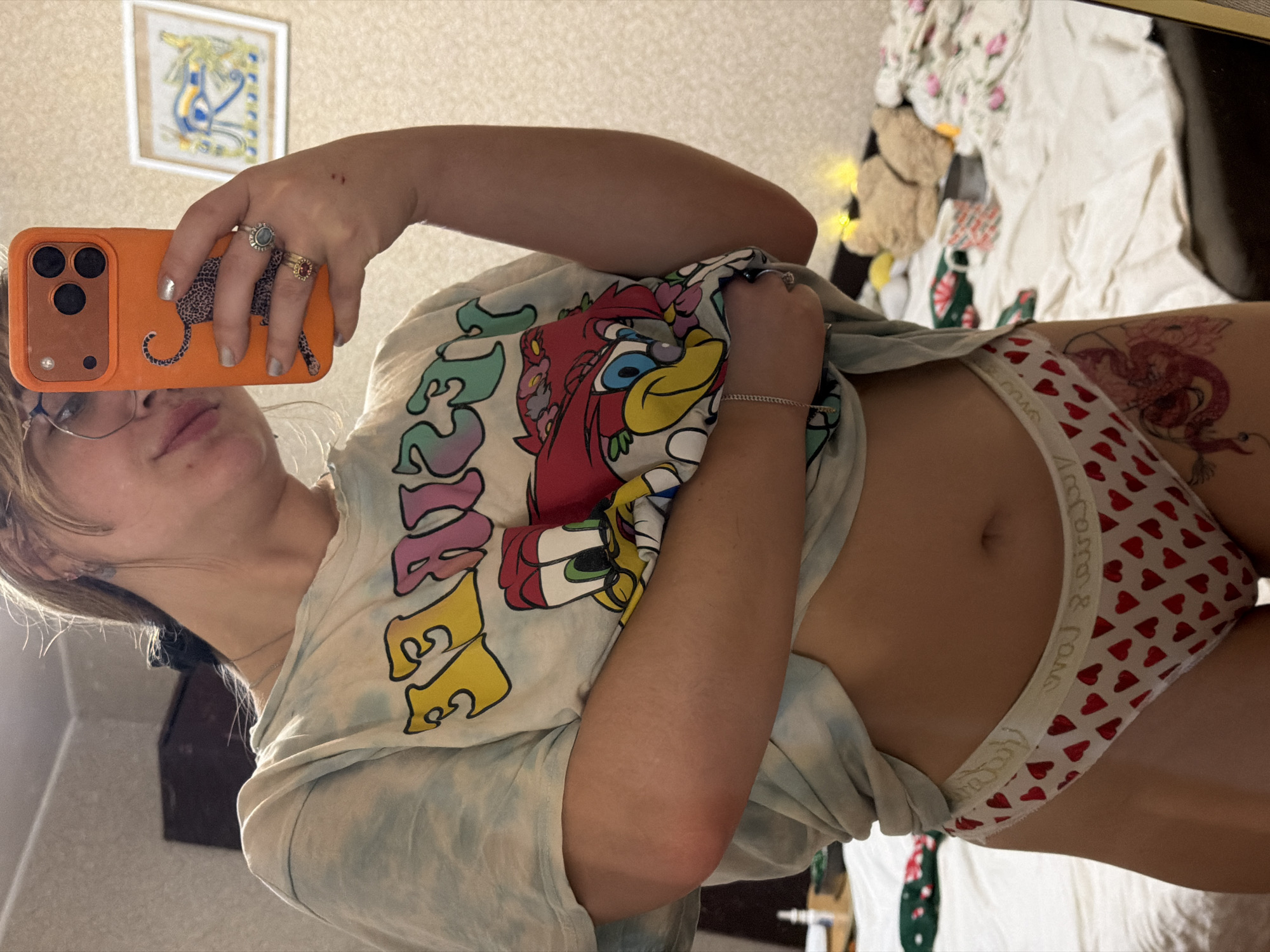 An indoor mirror selfie captures a person, likely female, from the chest down, rotated 90 degrees clockwise. The person wears a light blue tie-dye t-shirt featuring colorful cartoon characters, including what appears to be Daffy Duck and other Looney Tunes-style figures. The t-shirt is slightly lifted to reveal a bare midriff and white underwear adorned with small red hearts. The elastic band of the underwear has visible text, which appears reversed due to the mirror reflection, possibly reading "Arden & Love" or "Ariana & Love." A red tattoo, resembling a dragon, is visible on the person's upper right thigh/hip area.
The person is holding an orange phone with a black leopard print case in their right hand, wearing two rings on their fingers (one with a blue stone, another a gold band with a red detail) and light-colored nail polish. A thin bracelet is on their right wrist. Glasses are visible on their face, and their lips are slightly parted.
The background, reflected in the mirror, shows a light-colored wall with a framed picture featuring abstract yellow and blue art. Below, a cluttered surface, likely a bed or sofa, is covered with various textiles including a white blanket with pink flowers and another with a green and red holiday-themed pattern. A small brown stuffed animal, possibly a bear, and some decorative string lights are also visible. The setting appears to be a personal bedroom or living space.