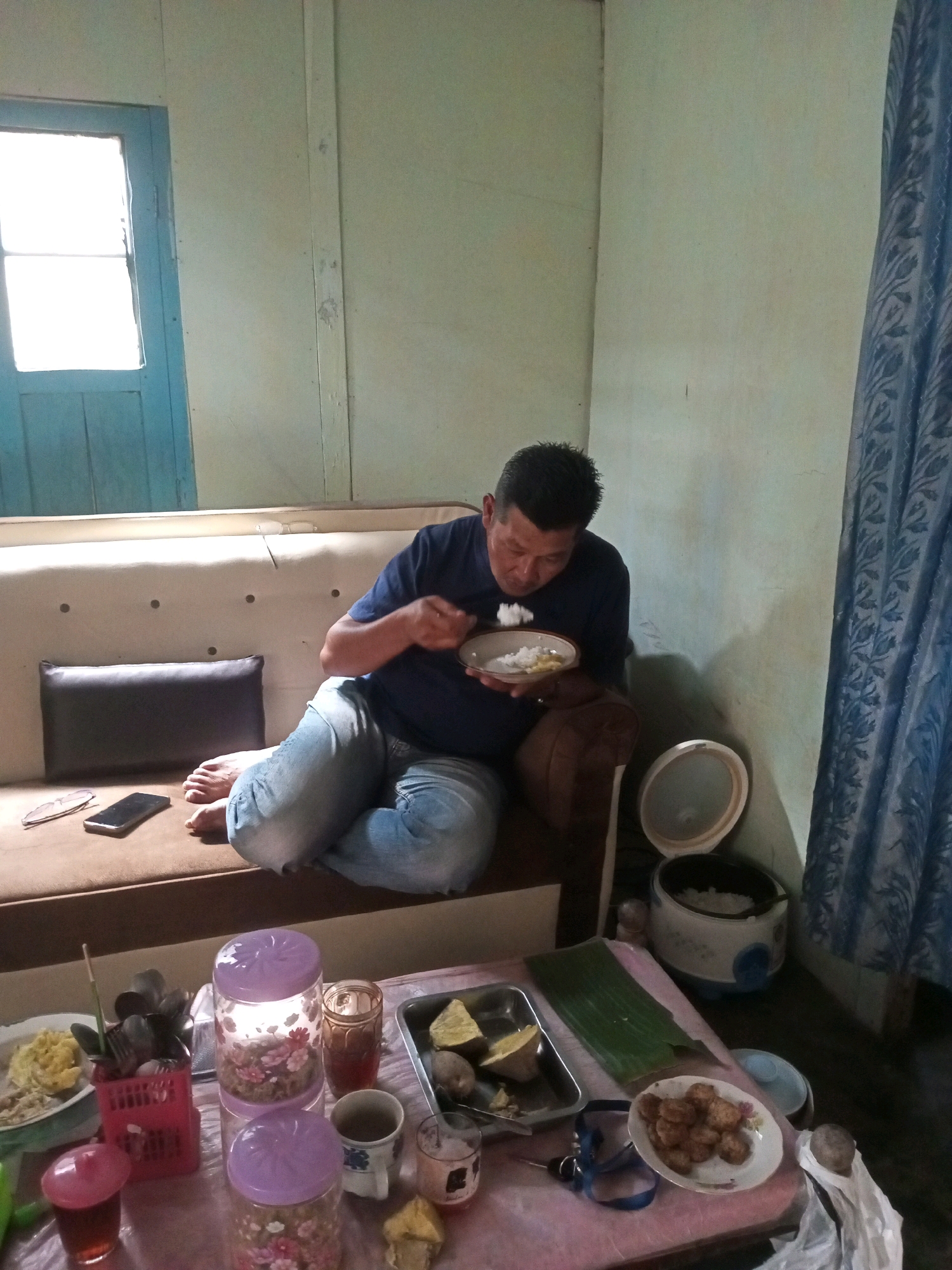 A man is sitting on a sofa in a room in Andongsili, Indonesia, eating from a plate. He is wearing a blue t-shirt and jeans. The sofa is light brown and has a dark brown cushion on it. There is a phone and a pair of glasses on the sofa next to the man. In front of the sofa, there is a table with food and drinks on it. There are also two jars with flowers on them. In the background, there is a blue curtain and a rice cooker. The room appears to be a living room.