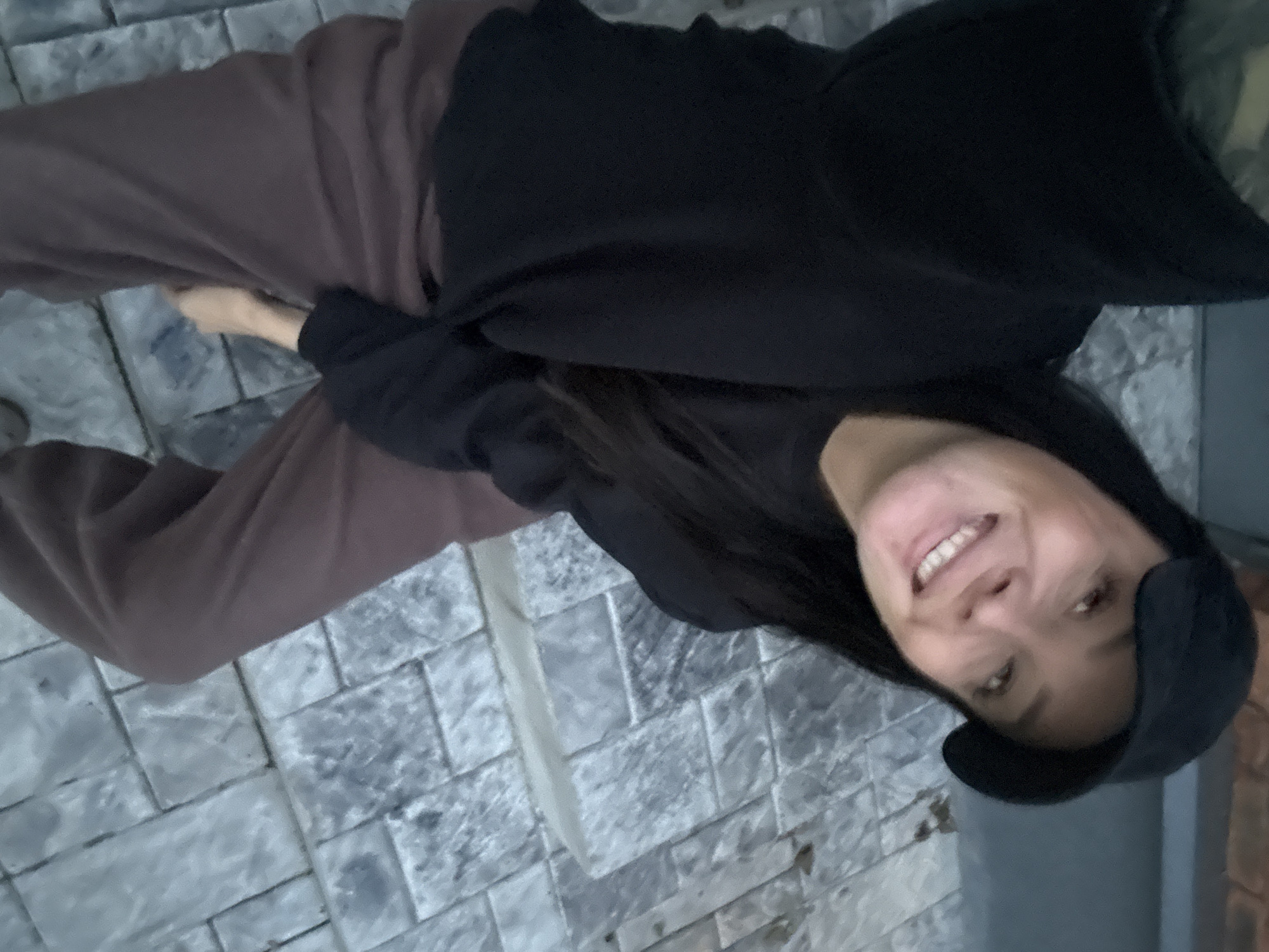 The image is a selfie taken by a woman wearing a black baseball cap, a black long-sleeved shirt, and brown pants. She is smiling and looking at the camera. The background appears to be a paved area with large, rectangular stones. The lighting suggests it is either early morning or late evening. The location is identified as Chaikovsky, Russia, but no specific landmarks or details confirm this.