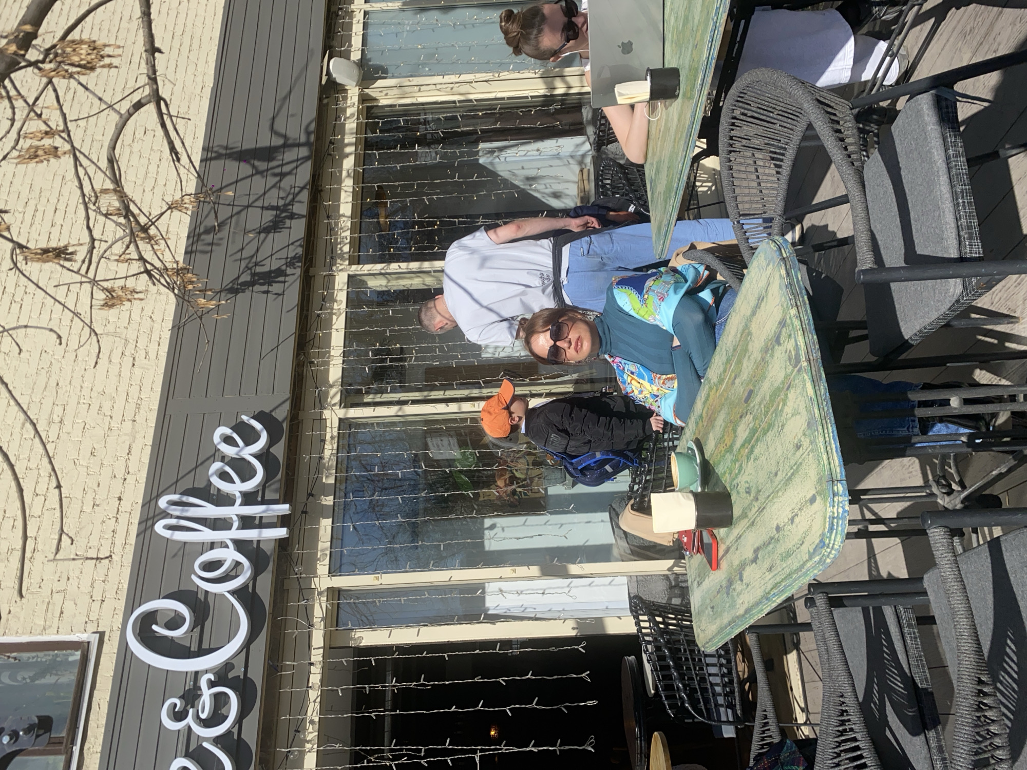 Here is a description of the image:
The image shows a group of people seated at tables outside a coffee shop. A sign reading "Coffee" is visible on the building. The scene includes men, women, and children sitting around the tables. The location may be Moscow, Russia. The outdoor area is decorated with string lights.