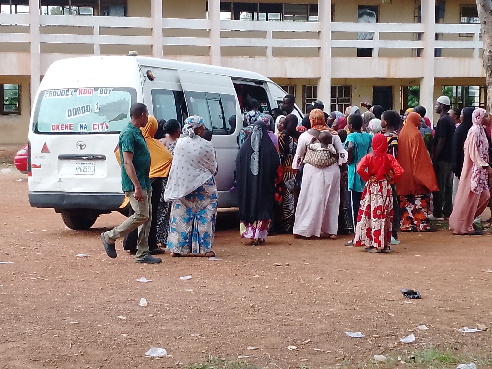 A crowded outdoor scene in Okene City features a white Toyota HiAce van parked beside a building with a white facade. The van's rear window displays the text "PROUDLY KOGI BOY" and "OKENE NA CITY." A group of people surrounds the van. Many of the people in the crowd are women wearing a variety of traditional African attire. One woman has a baby secured to her back with a carrier. A man in a green shirt and khaki pants walks to the left of the group. The ground is bare and scattered with trash. The background building has multiple windows and horizontal lines.