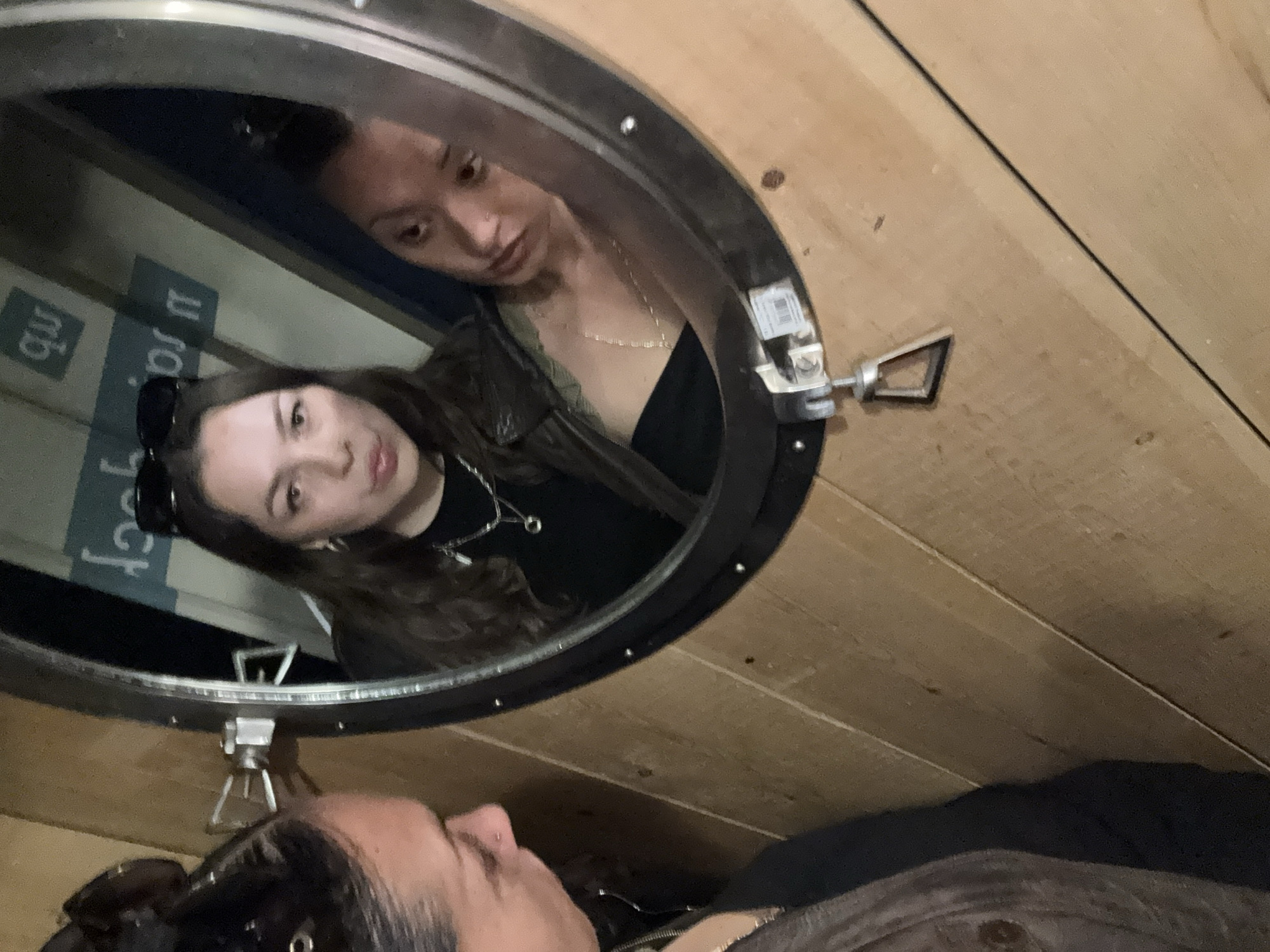 The image captures a selfie featuring two women within what appears to be a wooden interior. One woman is positioned in the foreground, likely holding the camera, with her face visible from the nose up, looking upwards into the lens. She has dark hair pulled back and wears a dark top.
Reflected in a prominent circular mirror, or porthole, is the second woman. She is seen from the chest up, with long dark hair, sunglasses resting on her head, and wearing a black top with a delicate necklace. Her gaze is directed slightly downwards and to her left within the reflection. Above her in the reflection, partially obscured, is another view of the first woman's face.
The circular mirror has a dark frame and features metal latches on its right side and bottom. The background behind the foreground woman and surrounding the mirror is composed of light-colored wooden planks.
In the reflection, beyond the second woman, some mirrored and upside-down text is visible on what appears to be a blue or dark surface. Partially legible characters include "mb" and "u ro". The overall setting suggests an enclosed, possibly unconventional space, like a cabin, a converted shipping container, or an art installation. The lighting is dim, suggesting an indoor or evening setting.