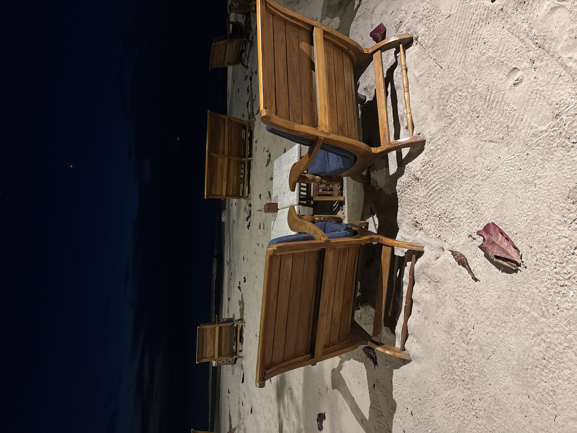The image depicts a peaceful night scene on a sandy beach, likely in Kuta Selatan, Indonesia. The perspective is from a slightly elevated angle looking down.
On the right side of the frame, light-colored sand covers the ground, showing clear texture, ripples, and some subtle indentations, possibly footprints. Several dried leaves, predominantly reddish-brown, are scattered across the sand.
Dominating the central and upper-right portions are multiple wooden chairs, appearing to be deck chairs or lounge chairs, crafted from polished, medium-toned wood. Each chair features a dark blue cushion. They are arranged on the sand, casting distinct, elongated shadows, indicating a strong light source from the left or above the frame, illuminating the beach area. Between some of the chairs, a small table with a light-colored surface is visible. All the chairs are unoccupied.
To the left, a large expanse of dark blue or black represents the night sky or the ocean, merging indistinctly in the low light. A few faint specks of light are visible in this dark area, suggesting distant stars or artificial lights. The scene conveys a quiet, tranquil atmosphere, typical of an uncrowded beach setting after dusk. No discernible text is visible in the image.