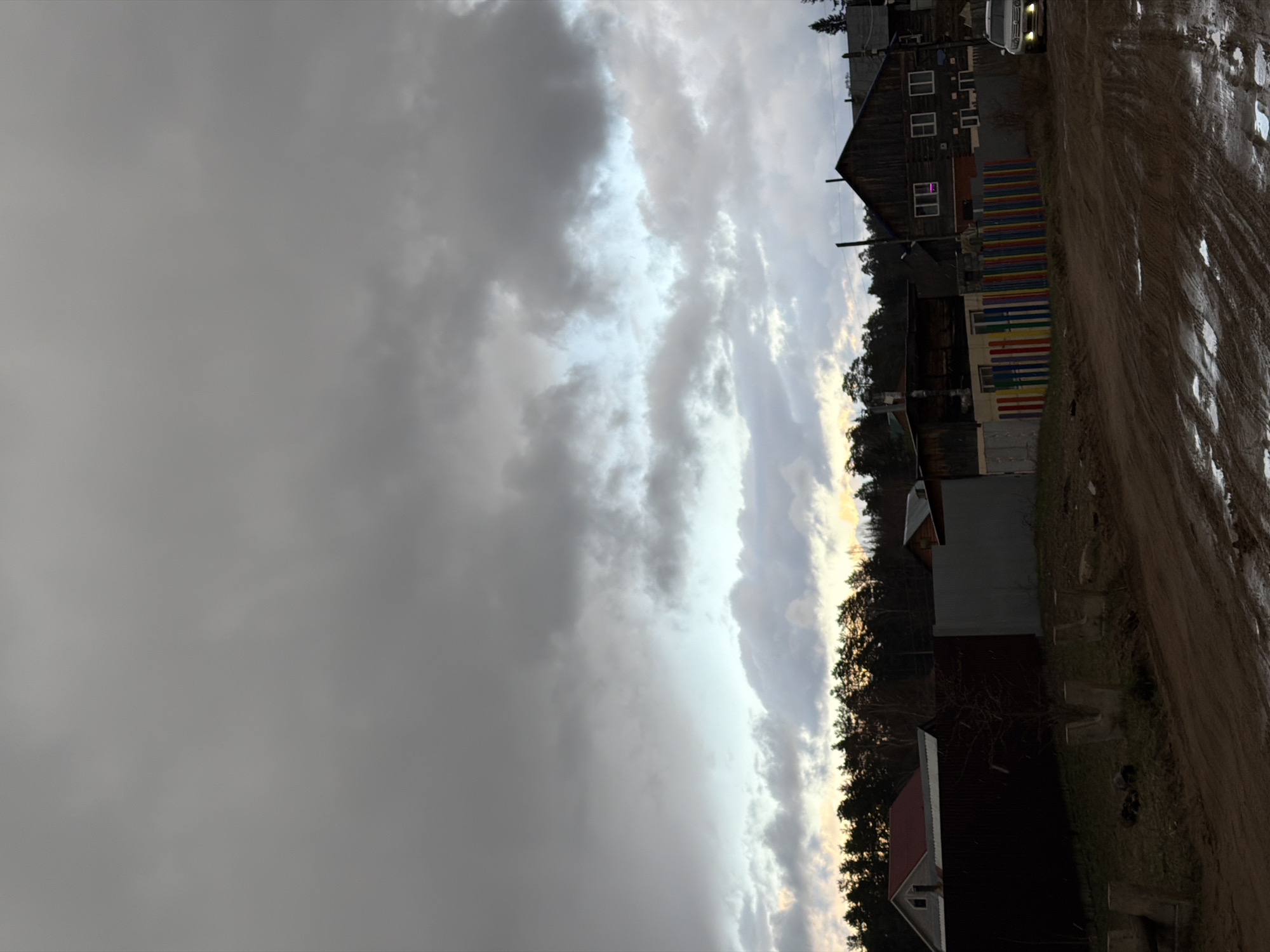 The image depicts a street in the city of Chaikovsky, Russia on a cloudy day. A muddy dirt road is on the right, with tire tracks visible. A row of houses lines the right side of the road, including a two-story house with rainbow-painted barriers. A vehicle is parked near the house. The sky is overcast.