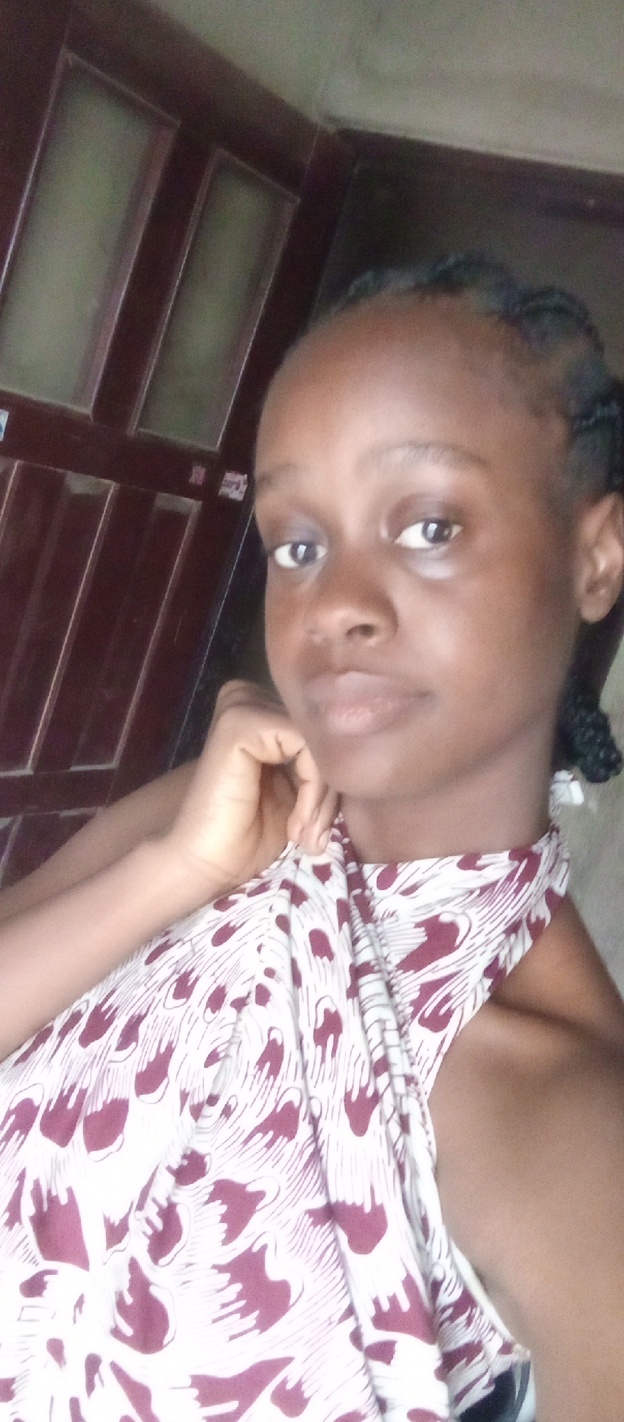 A close-up portrait features a young woman indoors in Akwanga, Nigeria. She has dark skin, dark eyes, and her hair is styled in intricate dark braids. She looks directly at the camera with a neutral, calm expression. She is wearing a light-colored, sleeveless top that features a distinctive dark red or maroon patterned design, which appears to be abstract feathers or swirling motifs. Her right hand is gently positioned near her chin or jawline. The background consists of dark wooden paneling or a door with several lighter, rectangular insets, indicating a simple interior setting. The lighting is soft and even, suggesting indoor ambient light during daylight hours. There is no visible text in the image.