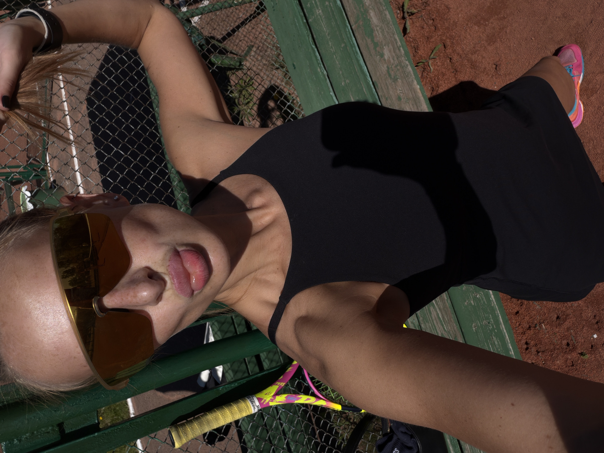 The image, taken in Krasnoyarsk, Russia, captures a person lying on a green wooden bench next to a tennis court. The subject, a woman with light-colored hair, is seen from a high-angle perspective, looking up towards the camera. She wears large, reflective golden-orange sunglasses and a black sleeveless top or dress. Her head is tilted to the side, and she playfully sticks out her tongue. One arm is raised, with her hand holding a strand of her hair, secured by a dark wristband.
The setting is an outdoor tennis court, featuring a reddish-brown clay surface and a green chain-link fence behind the bench. Bright sunlight creates strong, distinct shadows across the scene, indicating it is daytime. A colorful tennis racket, with a yellow, pink, and green grip, is partially visible, resting near the fence; the brand "Wilson" is clearly identifiable on its grip. A vibrant athletic shoe, primarily pink, blue, and orange, is visible on her right foot, resting on the clay court. Near the bottom left, a dark item displays partial white text resembling "BOSS." The overall scene suggests a casual and relaxed moment on the tennis court.
