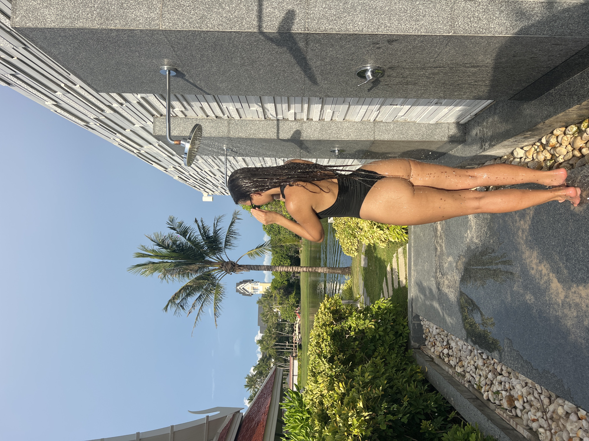 The image shows a woman showering outdoors in Choeng Thale, Thailand. She is wearing a black swimsuit and has long, dark braids. She appears to be rinsing her face or hair under the showerhead. The outdoor shower is attached to a wall constructed of dark grey granite tiles and light grey textured panels. Nearby is a palm tree, a lawn, a pond, and buildings. The sky is blue, suggesting it is daytime.