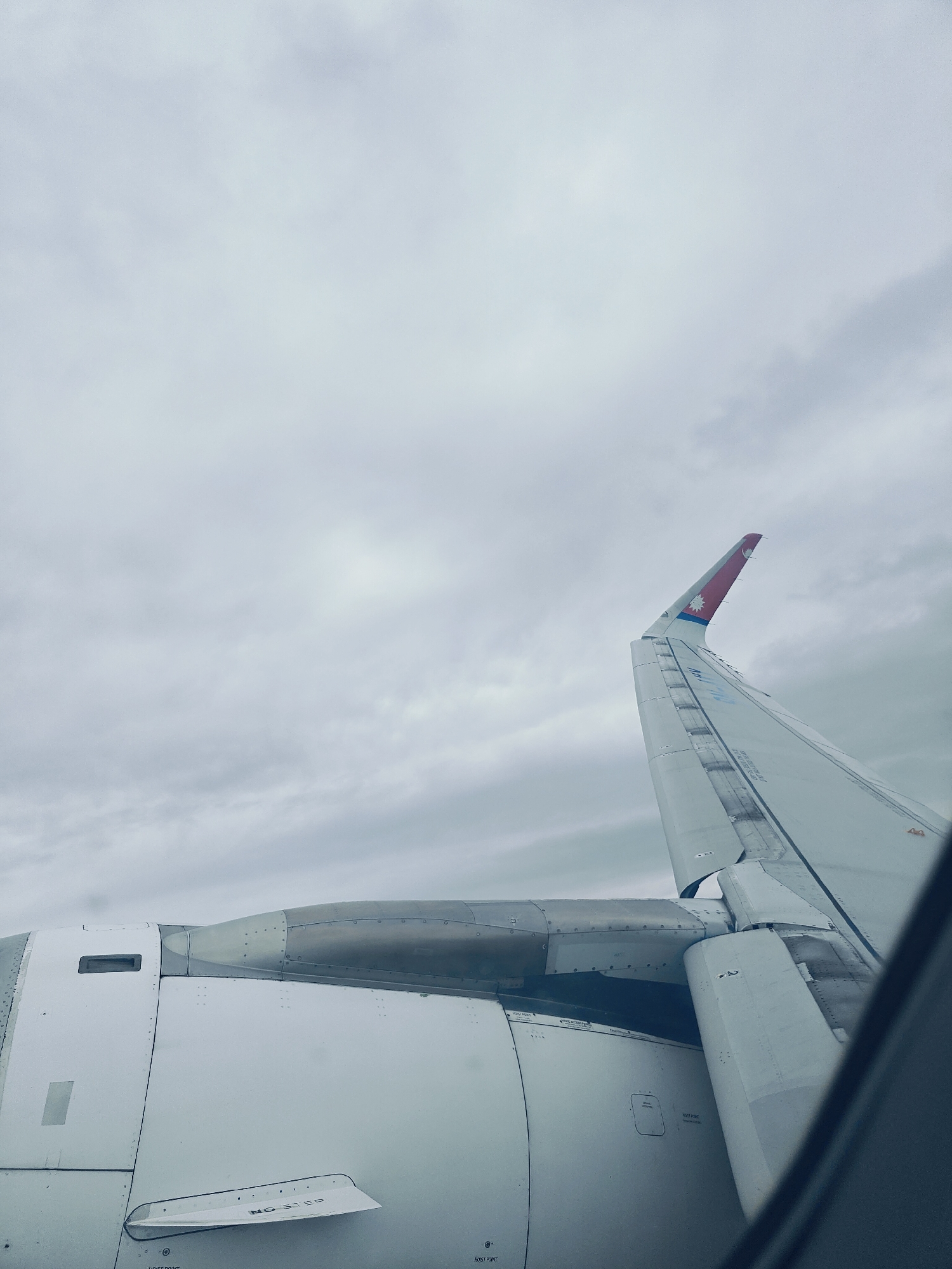 An aerial view from an airplane window shows a cloudy sky and the wing of the aircraft. The wing is white with red and blue markings on its tip, which resemble the flag of Nepal. In the foreground, the engine nacelle of the airplane is visible, also in white. The scene captures a moment during a flight, likely over or near Budhanilkantha, Nepal, given the aircraft's livery. There are no people or other identifiable objects in the image.
