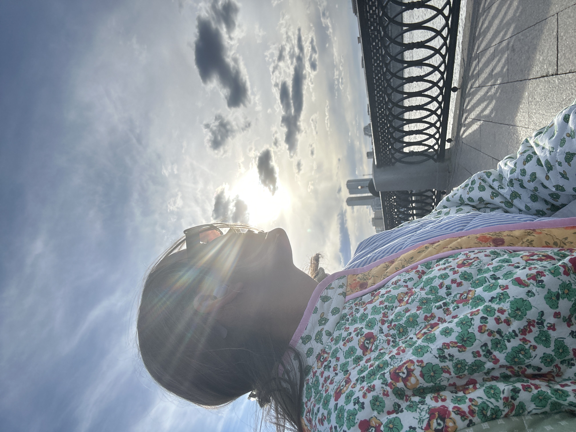 The media file captures a close-up, upward-looking shot of a person outdoors on a sunny, partly cloudy day in Moscow, Russia. The image is oriented vertically (originally rotated 90 degrees counter-clockwise), with the person's profile filling the lower-left foreground. They are largely silhouetted against the bright sun, which causes significant lens flare across the upper part of their face and head. Dark hair is visible, and what appear to be sunglasses are pushed up onto their head.
The person is wearing multiple layers of clothing or blankets. The most prominent is a white quilted fabric with a green leaf and red/orange floral pattern, suggesting a jacket or quilt. Below this, sections of yellow fabric and a blue-and-white striped fabric are visible.
The scene is dominated by a bright sky with scattered clouds, ranging from thin cirrus-like wisps to darker cumulus formations. The sun is intense, piercing through the clouds. To the right of the image, a decorative black metal railing (balustrade) with circular patterns is prominent. It casts intricate, elongated shadows onto a light-colored paved ground or promenade. In the far background, beyond the railing, several modern high-rise buildings are faintly visible, suggesting an urban setting.
The time of day appears to be mid-morning or afternoon due to the strong sunlight and distinct shadows. No visible text is present in the image.