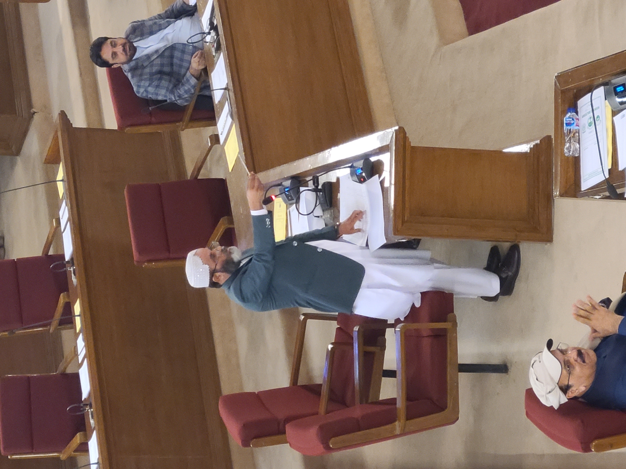 In a formal setting, possibly a legislative assembly in Quetta City Tehsil, Pakistan, a man wearing a white cap and a green blazer stands at a wooden podium, holding papers and speaking. The podium has microphones and other equipment. Several rows of maroon chairs are arranged behind the podium, and at least two other men are seated. One man to the right is wearing a white cap, and clapping his hands together. Another man at the far left is wearing a grey blazer. The interior appears to be a large room with wooden panels and furniture.