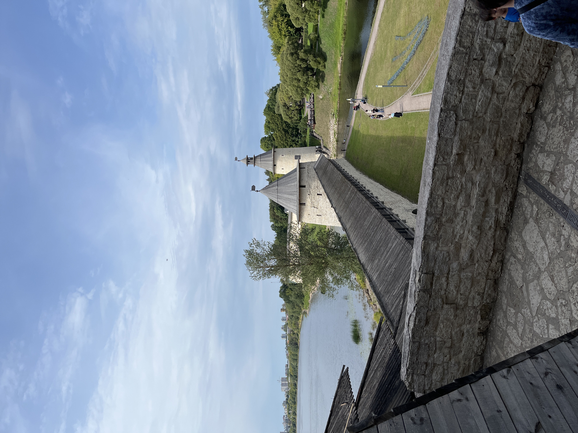 A wide, high-angle shot captures the Pskov Kremlin, a historic fortress in Pskov, Russia. The Kremlin's stone walls and conical-roofed towers dominate the mid-ground, with a wooden walkway running along the top of the wall. A body of water, likely a river, flows alongside the fortress, with a lush green bank dotted with trees. In the distance, the city skyline of Pskov is visible, characterized by buildings and cranes. A pathway meanders through the grass, with a few individuals strolling and admiring the scenery. The sky above is partly cloudy, with a light blue hue. The foreground features a stone wall and a dark wooden roof, suggesting the vantage point is from within the Kremlin's grounds.