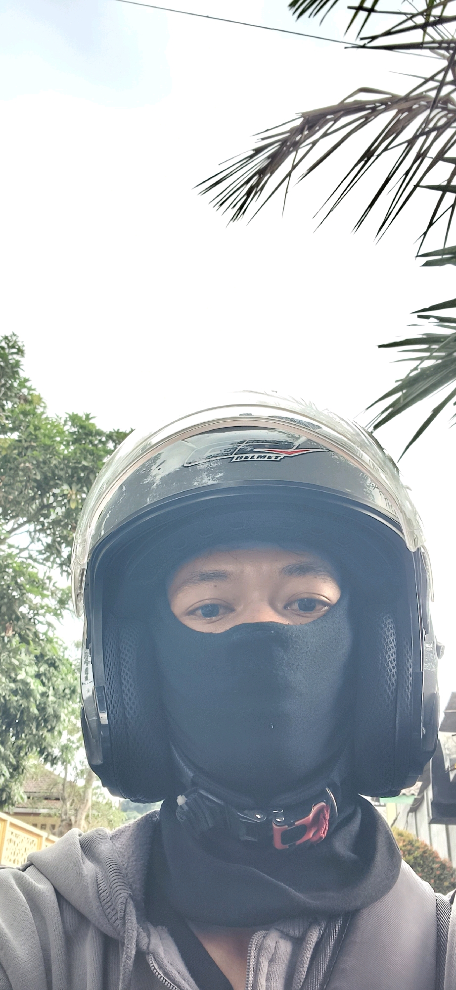 A close-up, low-angle selfie shows a person wearing a dark grey helmet and a black face covering. The helmet has a silver trim and a "FS Helmet" logo. The person's eyes are visible above the face covering, and they appear to be looking directly at the camera. A grey hooded jacket is worn over the person's shoulders. In the background, blurred green foliage from trees and a bright, overcast sky are visible. Some palm fronds hang into the frame from the top. The setting is outdoors, with hints of buildings and a fence in the distant background, suggesting an urban or suburban environment.
