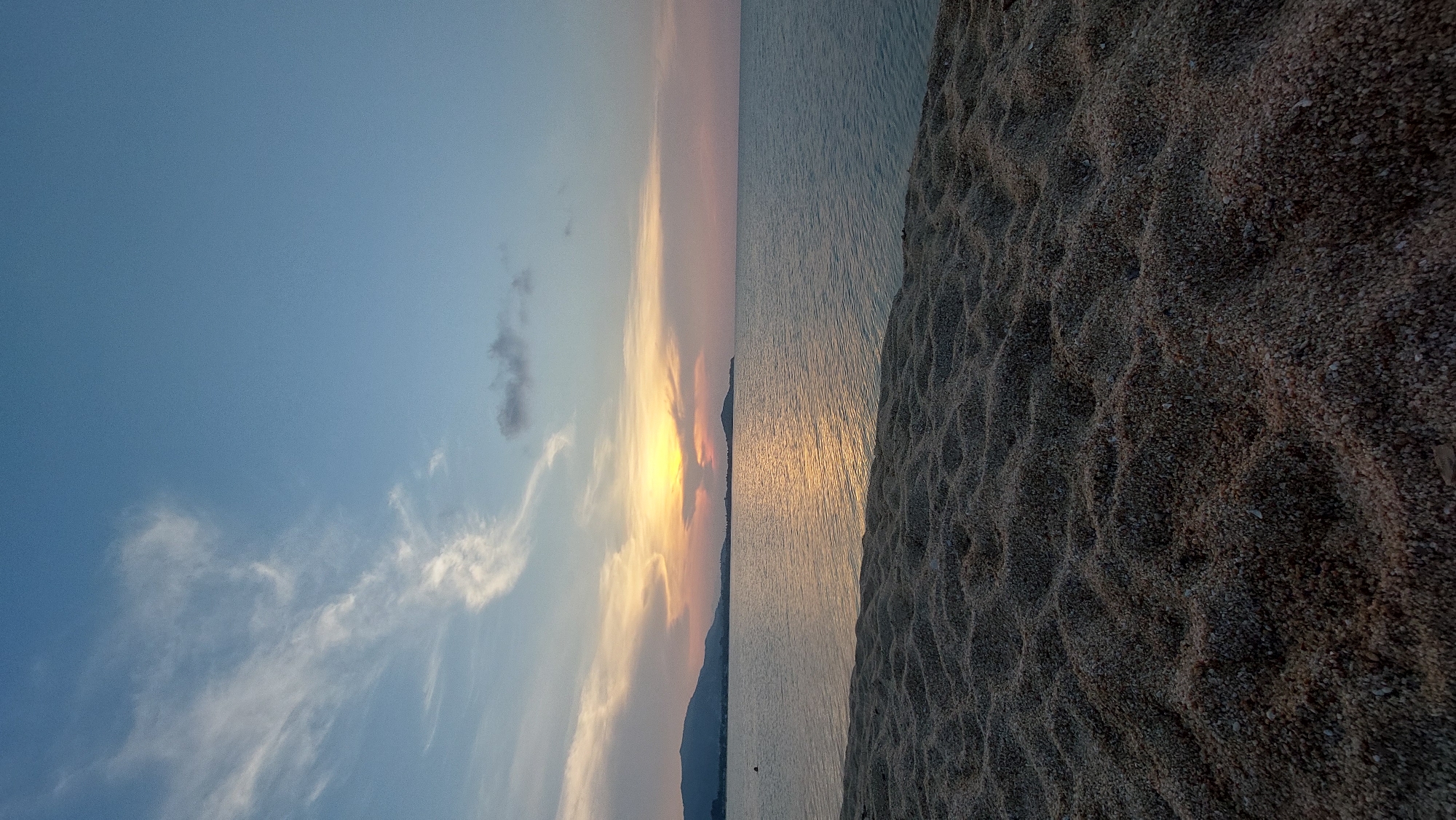 The image captures a serene sunset over a body of water, viewed from a pebble beach, likely in Baan Bo Phut, Thailand.
The upper portion of the frame features a expansive sky, transitioning from a clear, light blue at the top to a warm gradient of soft blues, pinks, oranges, and yellows near the horizon where the sun is setting. Scattered across the sky are thin, wispy white clouds, along with a few more textured, darker clouds.
The sun, a bright yellow-orange orb, is low on the horizon, partially obscured by distant landmasses. It casts a radiant, golden reflection across the tranquil surface of the water, creating a shimmering path of light. The water itself appears calm, showing subtle ripples and reflections of the sky's colors.
The foreground is dominated by the beach, composed of small, dark pebbles or coarse sand, rather than fine sand. The beach surface displays distinct, undulating patterns or ridges, suggesting the effects of receding waves or wind. This textured ground contrasts with the smooth expanse of the water and sky.
On the distant horizon, dark, silhouetted mountain ranges or landmasses stretch across the scene, providing a backdrop to the vibrant sunset. The time of day is clearly sunset, indicated by the low position of the sun and the warm, fading light. The weather appears clear and calm. There are no people, animals, or visible text present in the image, conveying a sense of peaceful solitude.