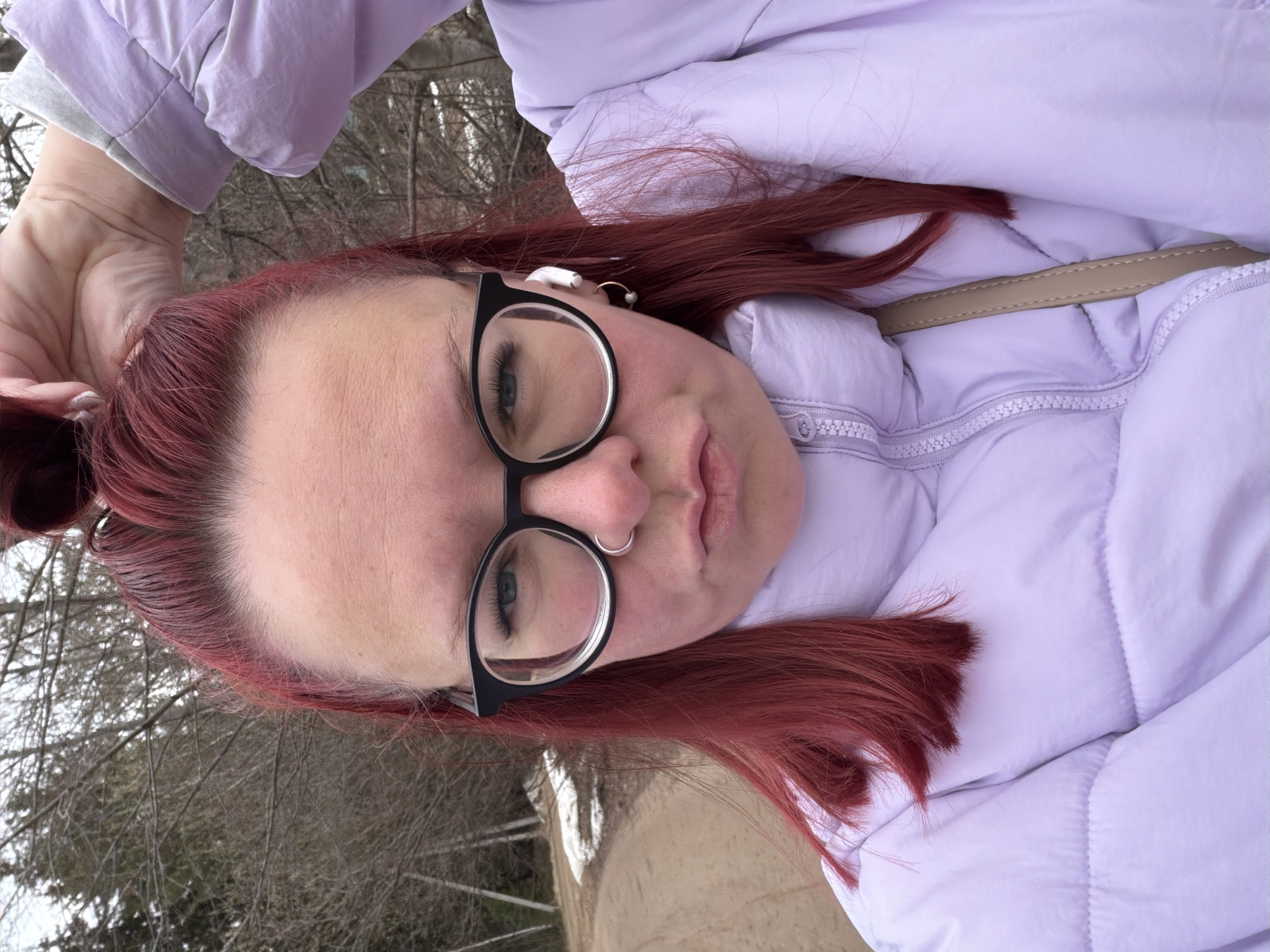 A woman with red hair, wearing black-rimmed glasses and a light purple puffer jacket, poses for a selfie. She is outdoors in a location, presumably Chaikovsky, Russia, with bare trees visible in the background. She is also wearing a nose ring. The woman has an earbud in her ear and appears to be holding her hair up. A beige purse strap is visible.