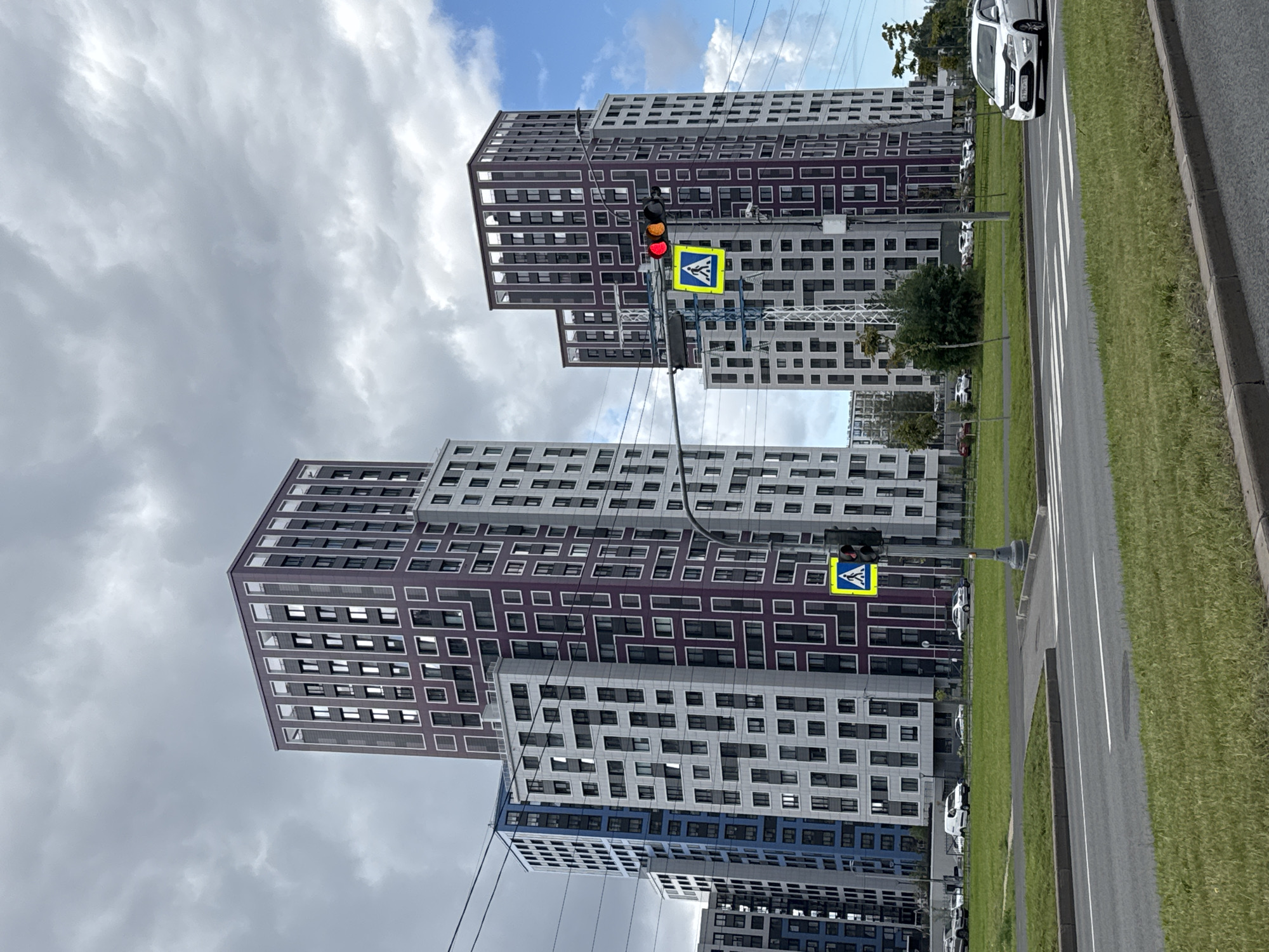 The image captures a wide view of a modern residential complex in Saint Petersburg, Russia, under a cloudy sky. In the foreground, a road with a pedestrian crossing is visible, with a white car driving across it from right to left. To the left of the road, a well-maintained grassy area stretches towards the buildings.
Dominating the scene are several tall, multi-story apartment buildings with distinctive architectural designs. These buildings feature a grid-like pattern of windows and balconies, with some sections highlighted in a deep purple color and others in white and light gray. The building on the far left has a blue facade.
A traffic light with red and amber lights illuminated is visible on a pole to the right of the central buildings, alongside a yellow pedestrian crossing sign. In the background, more buildings of similar design extend into the distance, and some trees are scattered across the landscape. The overall impression is one of urban development and modern living in Saint Petersburg.