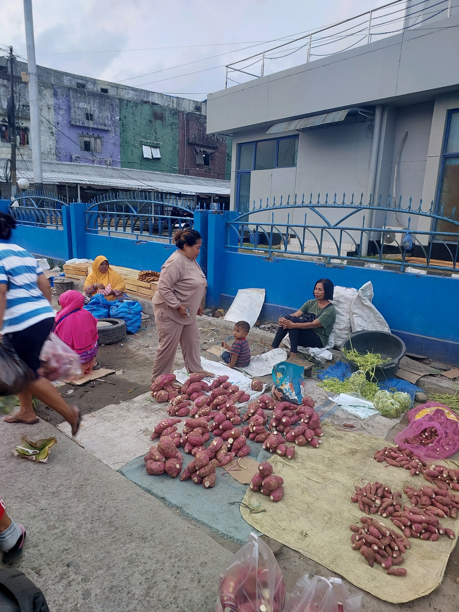 This is a street-level view of a market stall in Ambon, Indonesia, with a focus on the sale of sweet potatoes. In the foreground, a large pile of purple sweet potatoes is displayed on a light-colored mat. To the right, a woman wearing a green t-shirt and black leggings sits on a large sack, looking to her left. Next to her, a young child in a striped shirt sits on the ground. Behind them, a blue fence encloses a building with a grey facade and blue-trimmed windows. Further in the background, a multi-story building with a distressed, colorful facade (purple, green, and brown) stands against a cloudy sky. Several other individuals are visible in the middle ground: a woman in a beige outfit walks away from the camera, while another woman in a bright pink hijab and a woman in a yellow hijab are seated near some blue bags and what appear to be wooden planks. The overall scene depicts an outdoor market environment, likely in an urban area of Ambon.