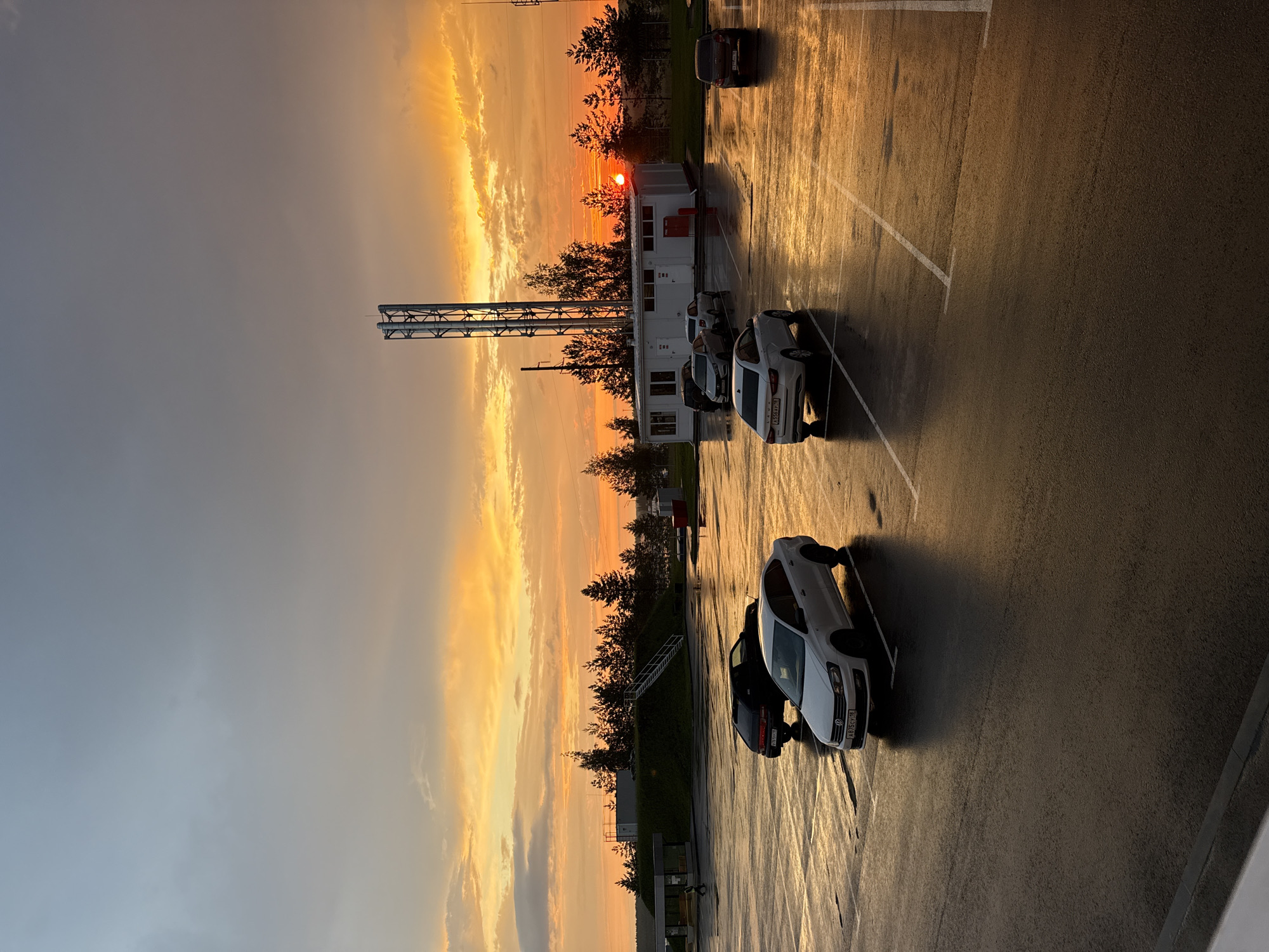 This image captures a parking lot in Sarapul, Russia, during a vibrant sunset. The sky is a dramatic mix of orange, yellow, and gray clouds, with the sun low on the horizon. The wet asphalt of the parking lot reflects the warm hues of the sunset. Several cars are parked, including a white Volkswagen Polo in the foreground, a white sedan behind it, a black sedan further back, and other vehicles obscured by distance. A white building with a tall industrial structure behind it stands on the left side of the scene, surrounded by trees. The overall atmosphere is serene, with the natural beauty of the sunset contrasting with the man-made structures.