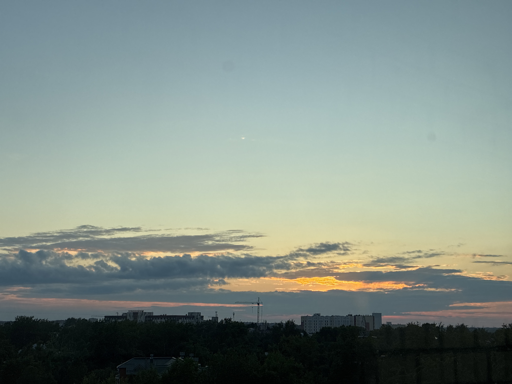 The image depicts a skyline at dusk or dawn in an urban environment. The sky is a gradient of pale blue to light yellow, with scattered clouds. The clouds are tinged with orange and pink hues, indicating the presence of the sun just below the horizon. A cityscape is visible in the distance, characterized by low-rise buildings and dense green foliage in the foreground, suggesting a residential or mixed-use area. A construction crane is visible among the buildings, implying ongoing development. The overall atmosphere is calm and serene, with the natural light of the sky providing the primary illumination. There are no people or animals visible in the frame.