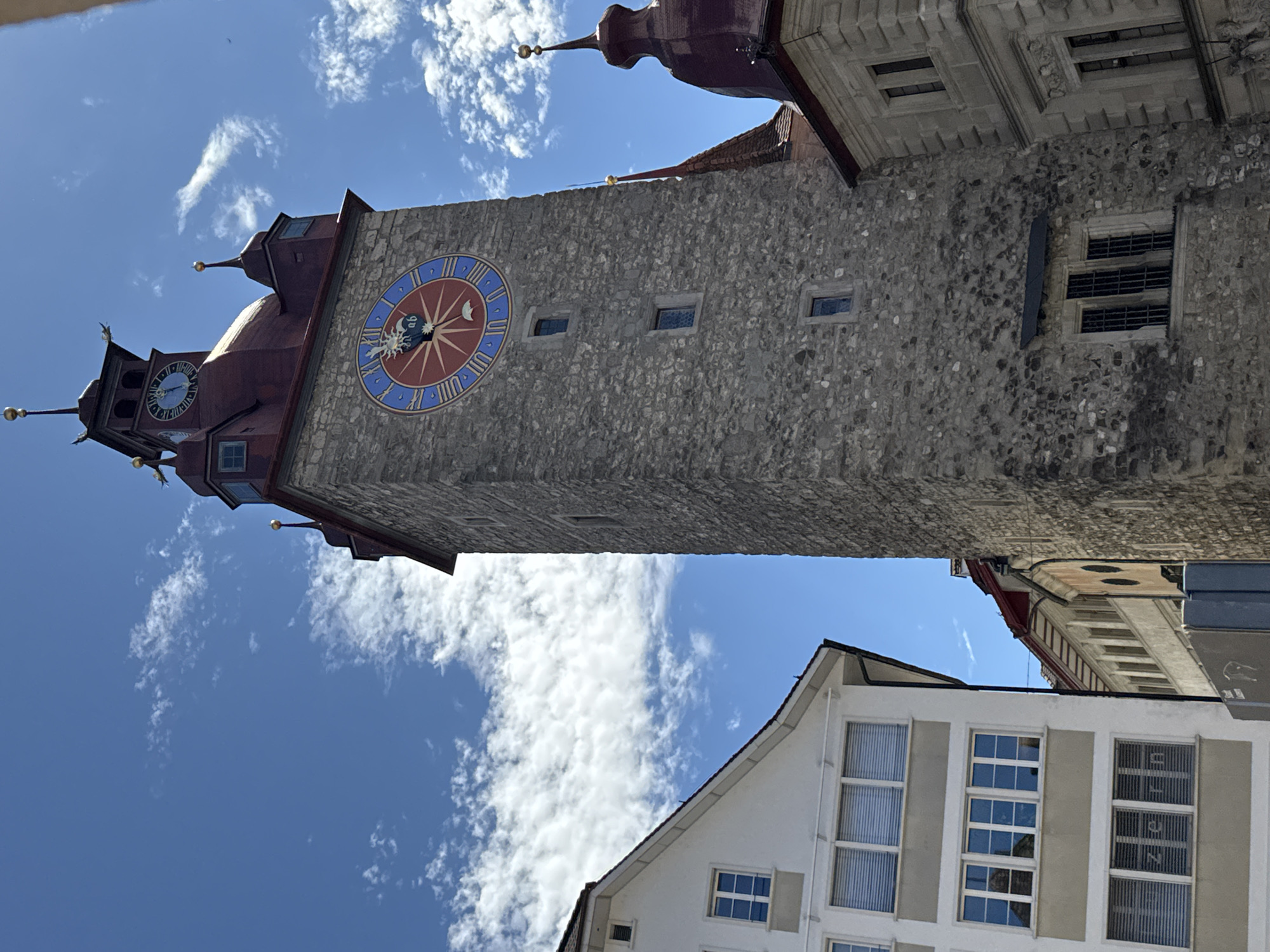 This is a low-angle, medium shot of the Zytglogge clock tower in Lucerne, Switzerland, on a sunny day with scattered clouds. The tower, made of stone, dominates the center of the frame with its large circular clock face featuring Roman numerals and a sun-shaped hour hand. The clock face is predominantly blue and red. The tower has a red conical roof with smaller turrets. To the left of the clock tower is a modern white building with multiple windows, some featuring blinds. To the right of the clock tower, partially visible, is another building with a red roof and architectural details. The sky is a clear blue with wispy white clouds. There are no people or animals visible in the image. The scene appears to be an urban setting focused on historical architecture.