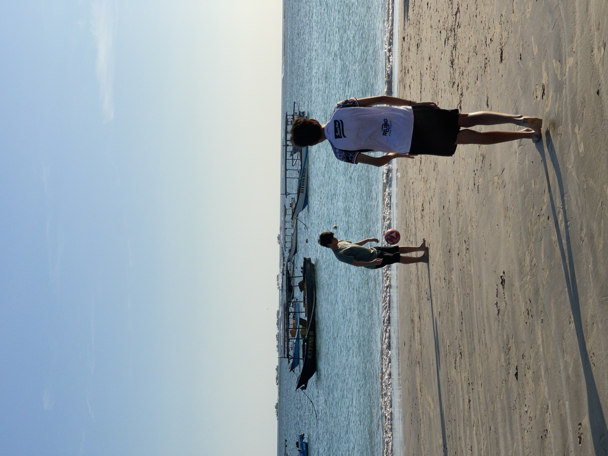 Two young males are playing soccer on the sandy shore of Kuta Beach, Indonesia. The sky is clear and blue, with the ocean stretching out to the horizon. Several boats are anchored in the calm waters. One male, wearing a green t-shirt and black shorts, is kicking a red and white soccer ball. The other male, facing away from the camera and wearing a white t-shirt with black trim and black shorts, is observing the game. The sun casts long shadows across the sand.