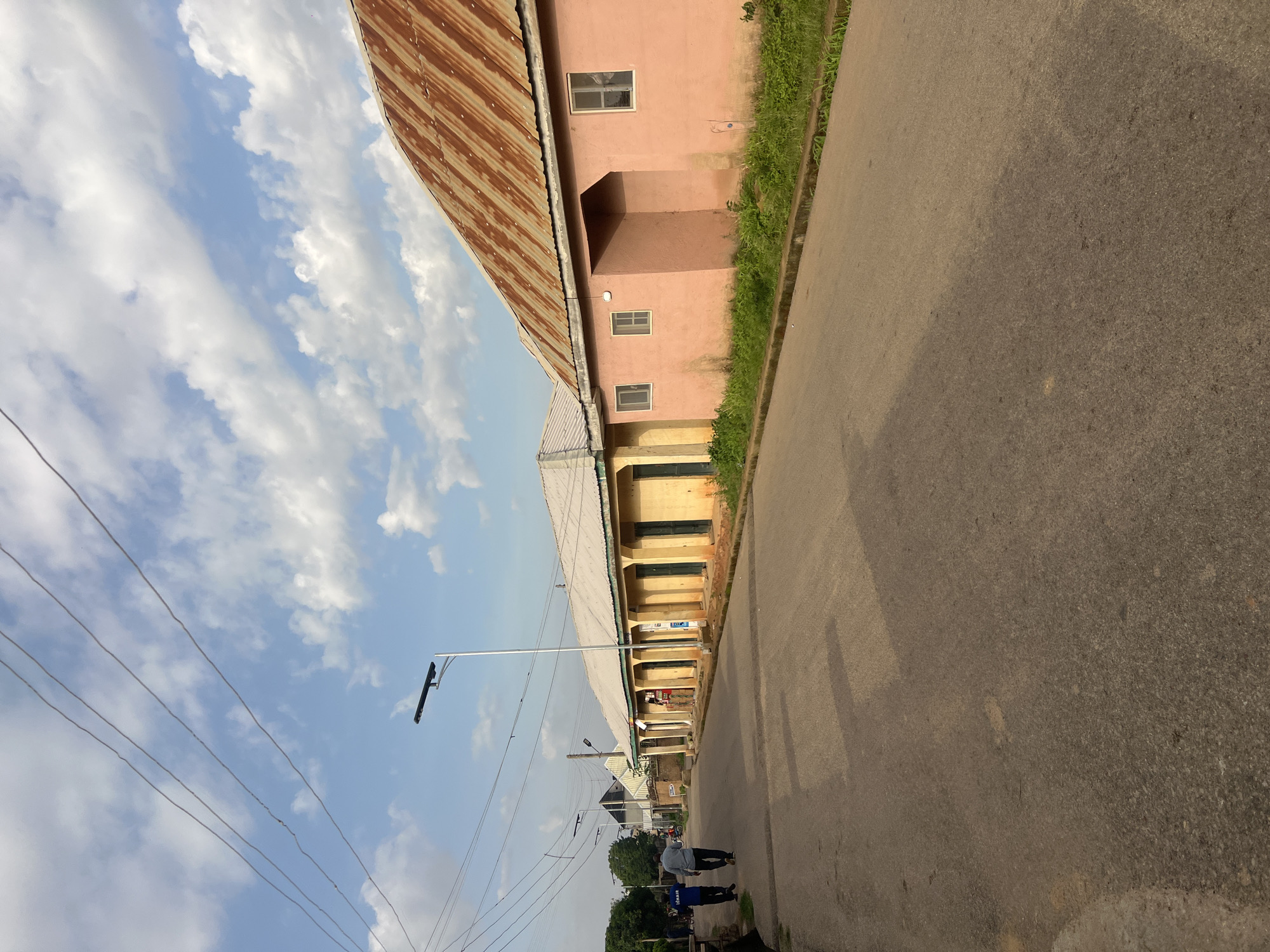 A straight, paved road runs diagonally across the image, bordered by a narrow strip of grass and a concrete curb. On the right side of the road, a row of single-story buildings with a distinctly pink facade and a rusted, corrugated iron roof is visible. The buildings have small, rectangular windows. Further down the street on the left, there is a longer, single-story structure with a light-colored roof and multiple arched doorways, painted in shades of yellow and green. Two men, dressed in casual clothing, are walking away from the camera on the left side of the road. Overhead, a clear blue sky is dotted with scattered white clouds, and electrical wires stretch across the scene, supported by utility poles. A modern street light is positioned near the center of the image. The overall impression is of a street scene in a residential or commercial area, likely in a climate with warm weather, as suggested by the building styles and the presence of greenery. The architecture and the informal arrangement of buildings suggest a location in Nigeria, potentially within the city of Jos.