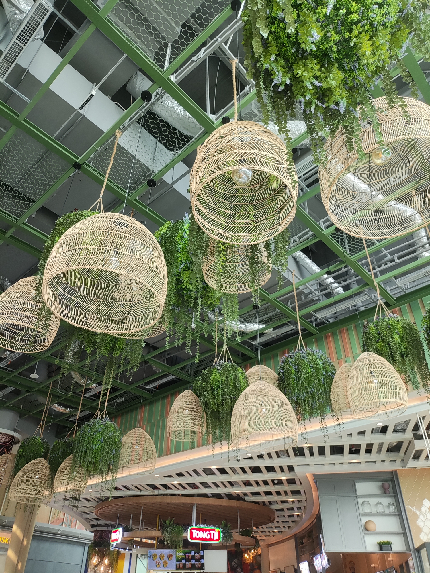 The image shows a view looking up at the ceiling of an indoor space, likely a restaurant or shopping mall, decorated with hanging wicker lamps and artificial plants. The ceiling structure is made of green metal beams and mesh. Several large, woven wicker pendant lamps hang at various heights, some illuminated by exposed bulbs within. Draping artificial greenery, with small purple accents on some, cascades from above and around the lamps, creating a natural, bohemian aesthetic. In the background, glimpses of the underlying ceiling infrastructure, including ventilation ducts and pipes, are visible. At the bottom of the image, a sign reading "TONG TI" is visible, suggesting a food establishment. The overall scene conveys a modern, eco-conscious interior design. There are no people visible in the image.