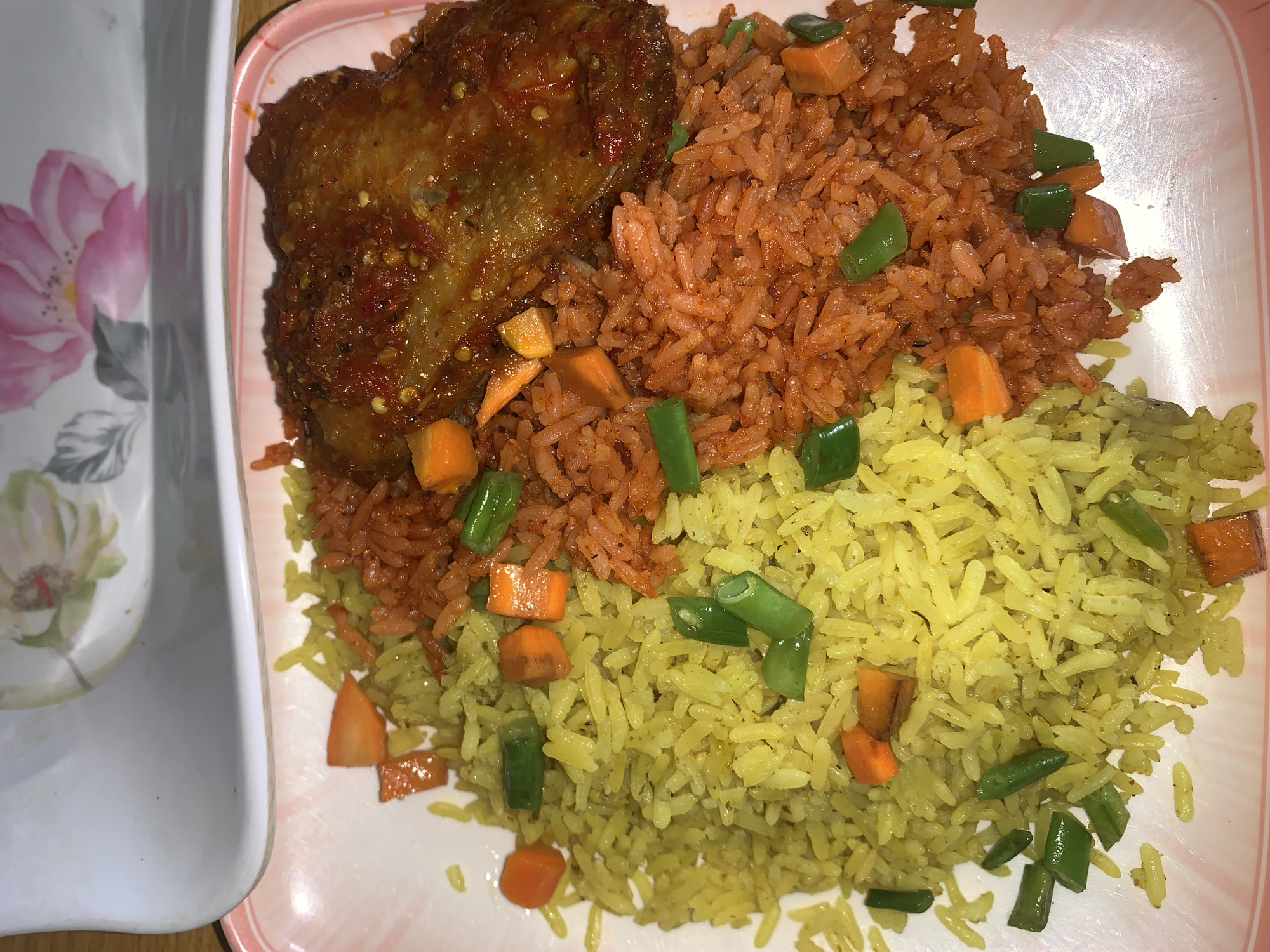 The image shows a plate of food on a table. The plate contains two types of rice, one red and one yellow, mixed with diced carrots and green beans. A piece of cooked meat covered in a red sauce sits on top of the red rice. The plate has a pink and white rim, and a separate white plate with a floral pattern is partially visible to the left. The location is Kabong, Nigeria.
