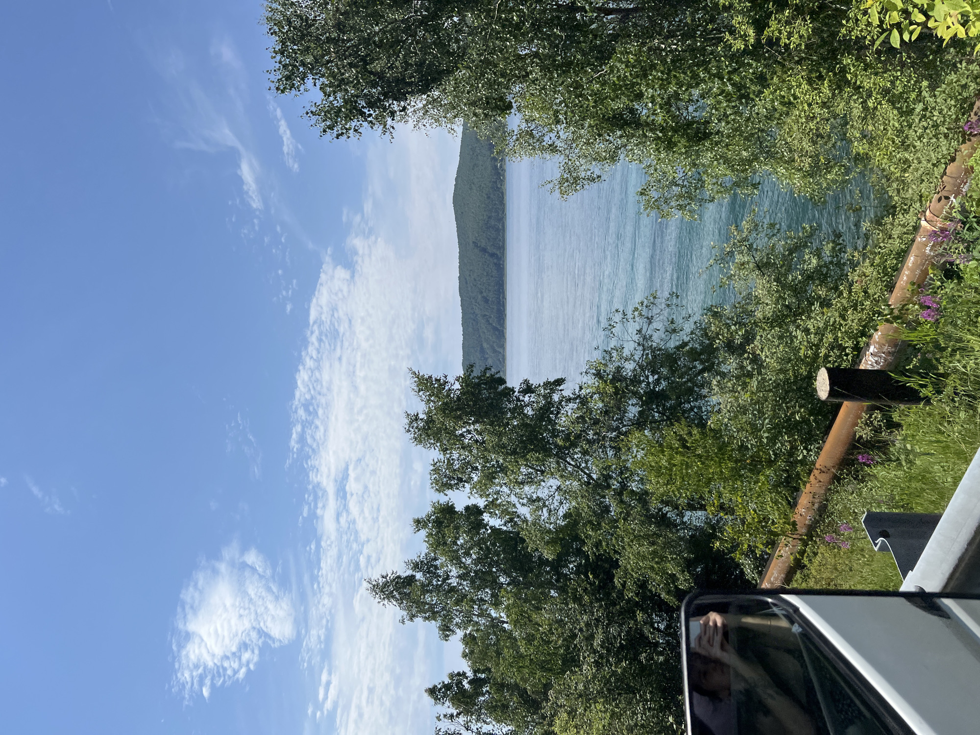 The image depicts a scenic view of a large body of water, likely a lake or sea, with a forested hill in the distance under a bright blue sky with scattered white clouds. Lush green trees and foliage frame the foreground and the water, with some rust-colored pipes and a wooden post visible near the bottom of the frame. In the lower left corner, a reflection in a vehicle's side mirror shows a person holding a phone, suggesting the photo was taken from inside a moving vehicle. The environment appears to be natural and possibly rural.