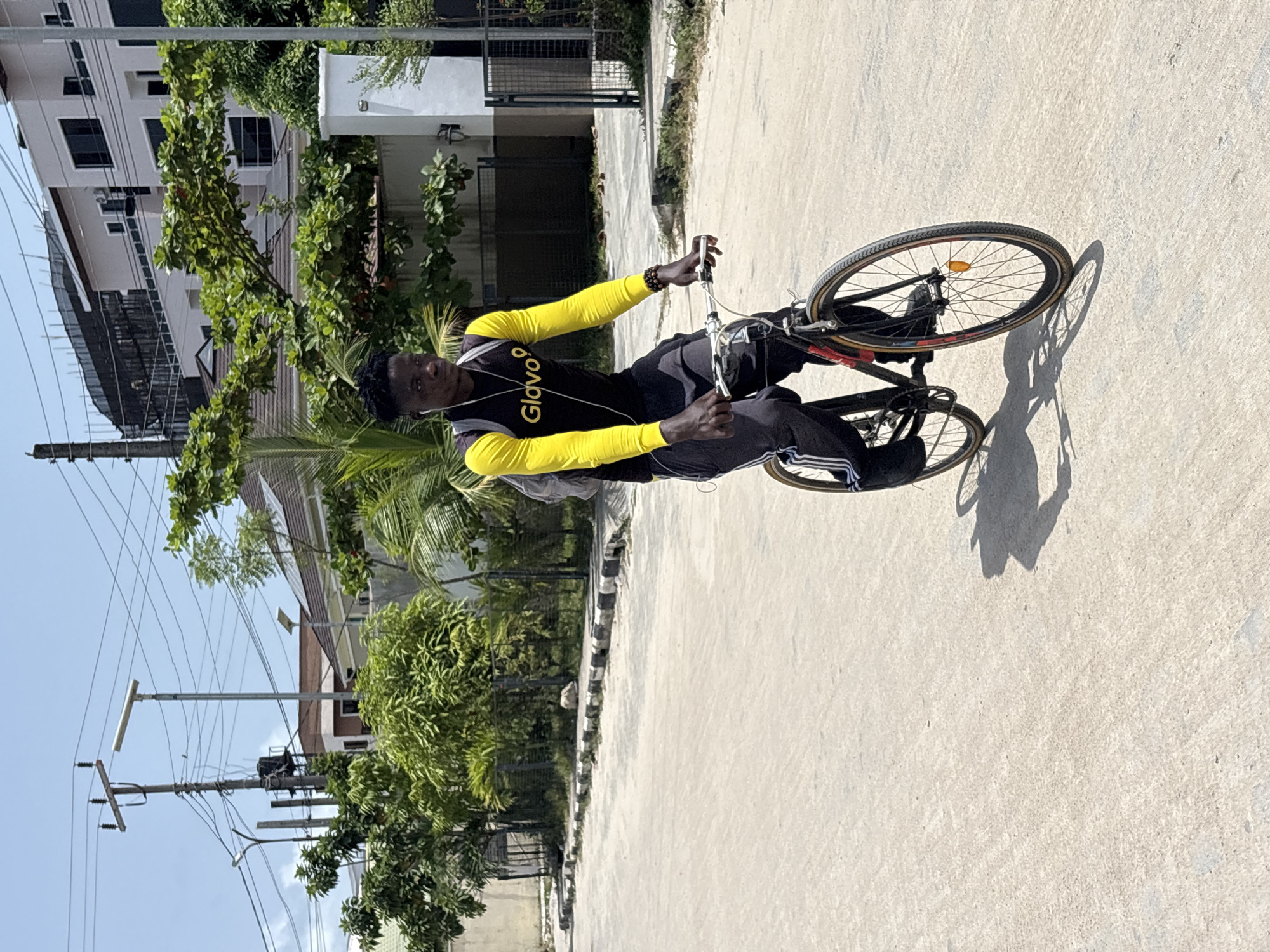 The image captures a young Black man standing next to a bicycle on a paved road under bright sunlight. He is positioned on the right side of the frame, looking directly at the camera with a neutral expression. He wears a black short-sleeved shirt with the word "Glovo" prominently displayed in yellow text on the chest, paired with long yellow sleeves underneath. He also wears black track pants with white stripes down the sides and black sneakers. White earphones are visible. He holds the handlebars of a dark-colored bicycle, with its front wheel slightly lifted off the ground.
Strong shadows of the man and bicycle are cast on the light-colored, possibly concrete, road, indicating a sunny day. In the background, to the left, several multi-story buildings with light-colored facades and dark windows are visible amidst abundant lush green trees and bushes, including palm fronds, suggesting a tropical environment. Further left and towards the bottom, multiple utility poles with numerous electrical wires crisscross against a clear blue sky. A curb with black and white stripes runs alongside the road in the mid-ground. The scene depicts a daytime setting in what appears to be an urban or semi-urban area, consistent with Igboefon, Nigeria.