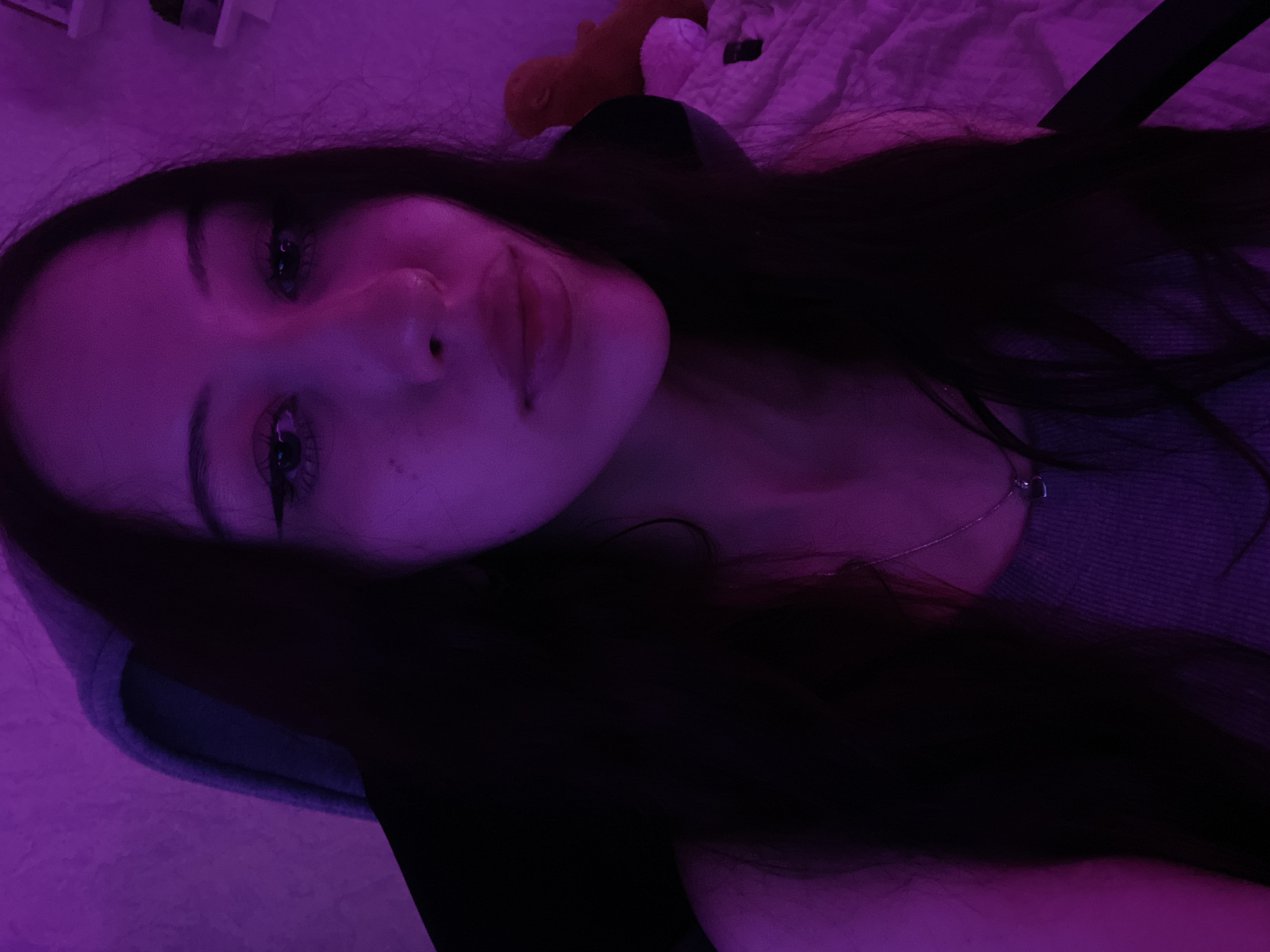 The image shows a close-up, chest-up selfie of a young woman with long, dark hair. She is wearing a grey tank top and a silver necklace with a small pendant. The scene is illuminated by purple light, creating a distinct aesthetic. The background includes a bed with a white blanket and a small orange stuffed toy. There is also a hint of a shelving unit on the right side of the image. The woman has makeup on, including eyeliner and mascara. The overall environment appears to be a bedroom. There is no discernible information about the location being in Votkinsk, Russia.