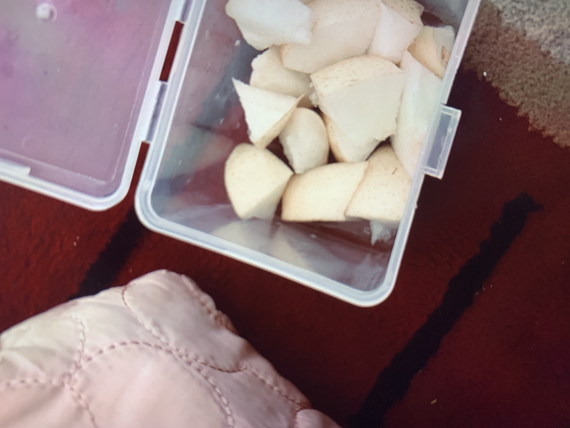 A close-up, overhead shot shows a clear plastic container filled with chopped pears. The container is slightly open, with its lid pushed back, revealing the white, segmented fruit. To the left of the container is a soft, light pink, quilted object, possibly a bag or cushion. The background is a dark red, textured surface, possibly a tablecloth or rug. The overall scene appears to be indoors, likely on a table, with no clear indication of a specific location within Ciamis, Indonesia.