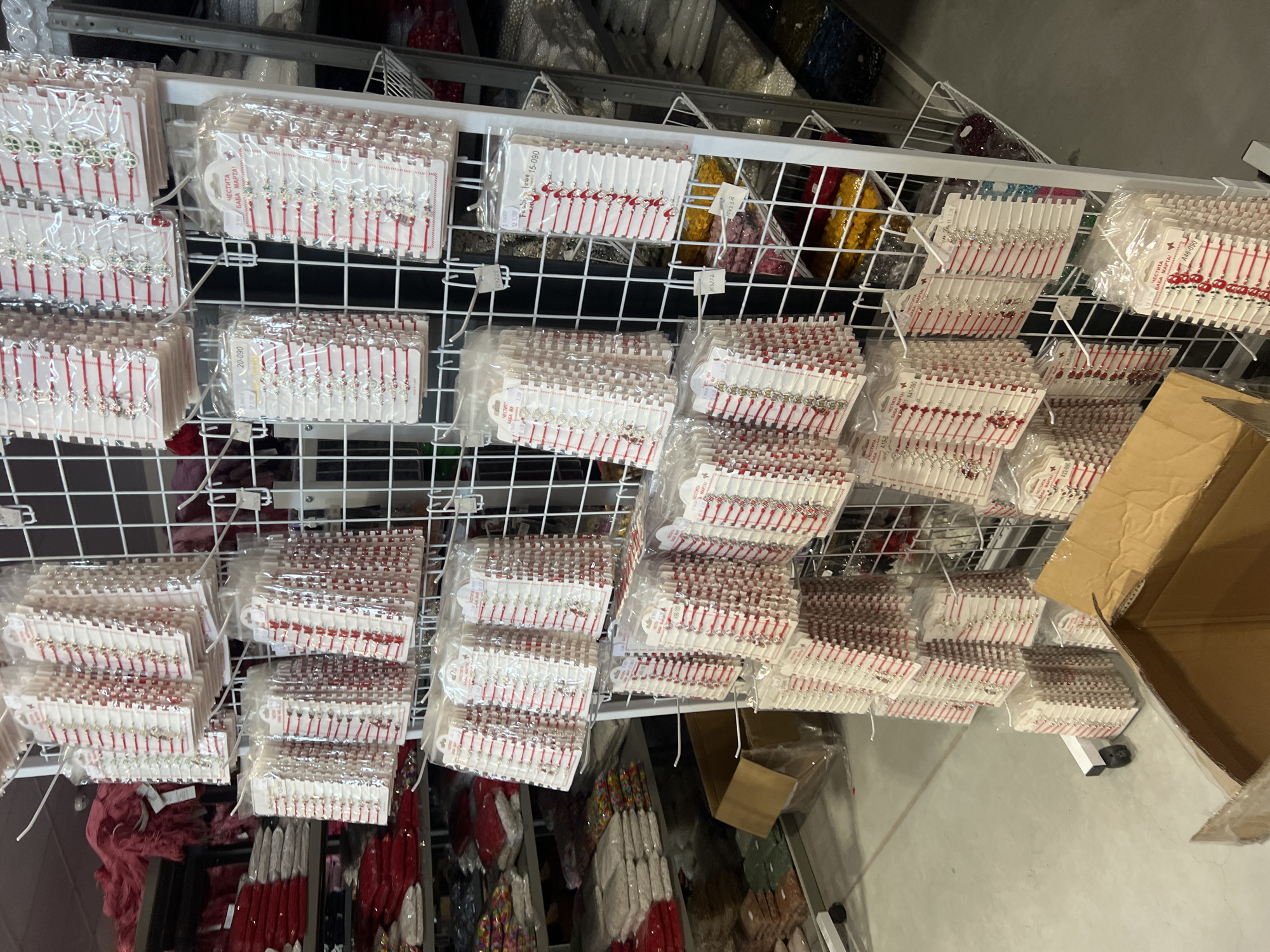 The image shows a commercial setting, possibly a retail store or storage area in Burgas, Bulgaria. Numerous packets of "Martenitsi" bracelets, traditional Bulgarian charms worn in March, are displayed on a white wire rack. These bracelets consist of red and white threads, often with small charms attached. The packets are stacked in neat rows, suggesting organized storage or display for sale. The packaging is transparent, showing the bracelets inside. The background reveals more inventory and storage items. The lighting is artificial and bright.