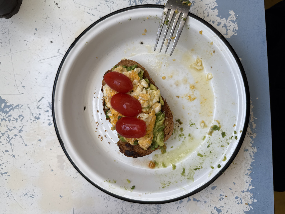 A partially eaten piece of toast sits on a white plate with a black rim, accompanied by a fork. The toast is topped with mashed avocado, scrambled eggs, and three small, red tomatoes. Traces of avocado and egg residue smear the plate's surface. The plate rests on a table with a mottled, light-colored pattern. Part of the image is obstructed by black color.