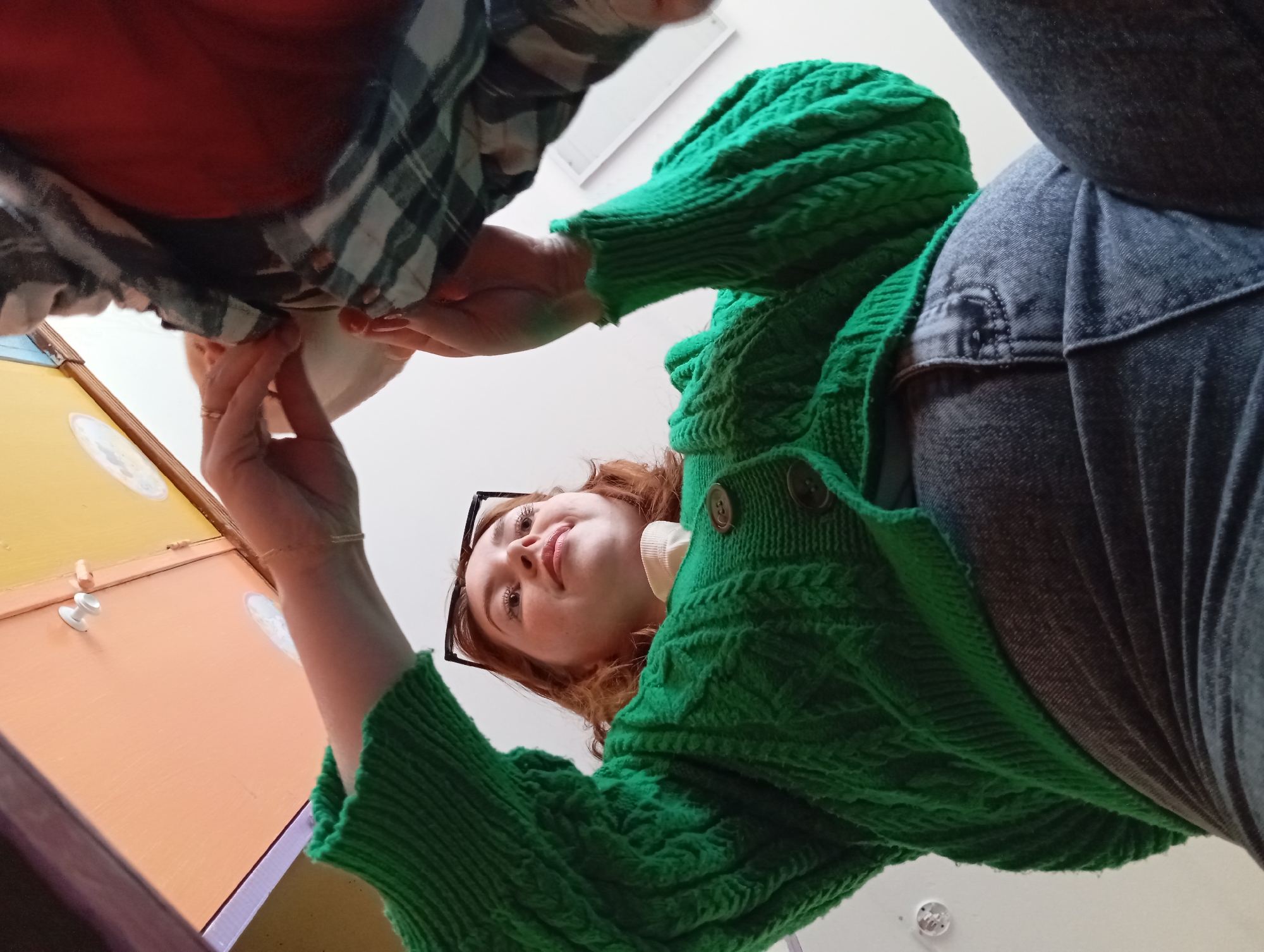 A low-angle shot features a young woman with red hair, wearing a green knitted cardigan and black-framed glasses, looking down at the camera. The perspective indicates she is standing above the viewer. Her hands are holding a white object, possibly a plate or bowl, which is also being held by another person partially visible in the upper left corner of the frame. This second individual is wearing a plaid shirt. The background includes a cabinet or drawer set with an orange face and white knobs. The setting appears to be indoors.