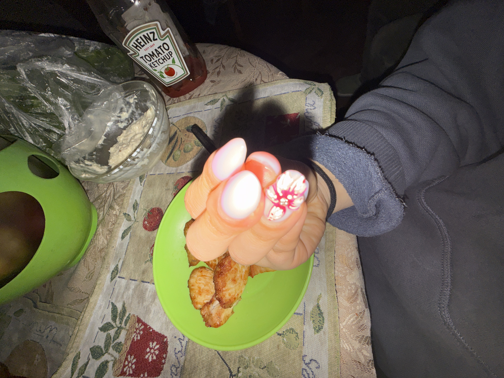 The scene depicts a person's hand holding a green plate with fried food. The person is wearing a gray sweatshirt and has a black bracelet on their wrist. Their nails are painted with a white and pink floral design on one finger and an ombre effect on the others. The plate sits on a tablecloth with a floral pattern. To the left of the plate is a green container with a brown object inside, a clear bowl with food, and a bottle of Heinz tomato ketchup. The image was captured in an indoor environment. There is no indication of this image being from the city of Sofia, Bulgaria.