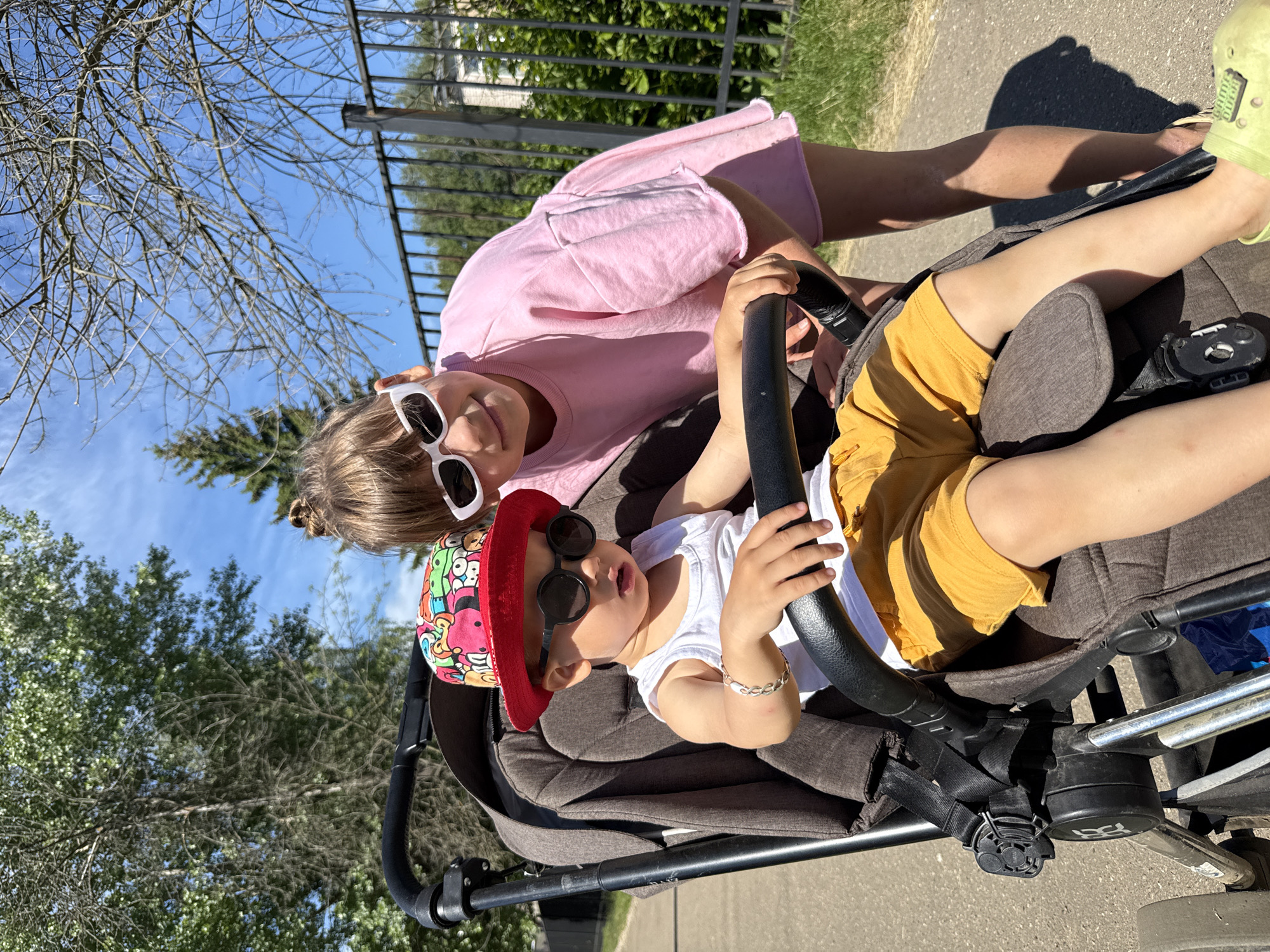 Two children, a boy and a girl, are in an outdoor setting, likely in the city of Rusanovo, Russia, on a sunny day. The boy, sitting in a stroller, wears a white tank top, yellow shorts, a red bucket hat with a colorful pattern, and black sunglasses. He has a silver bracelet on his left wrist and is holding onto the stroller's handlebar. The girl, standing behind the stroller and leaning over, wears a pink t-shirt and white sunglasses. She has her hands on the stroller's handlebar, interacting with the boy. The stroller itself is dark brown and appears to be in use. The background includes green trees, branches of a leafless tree, a black metal fence, and a paved path. The sky is blue with some wispy clouds.