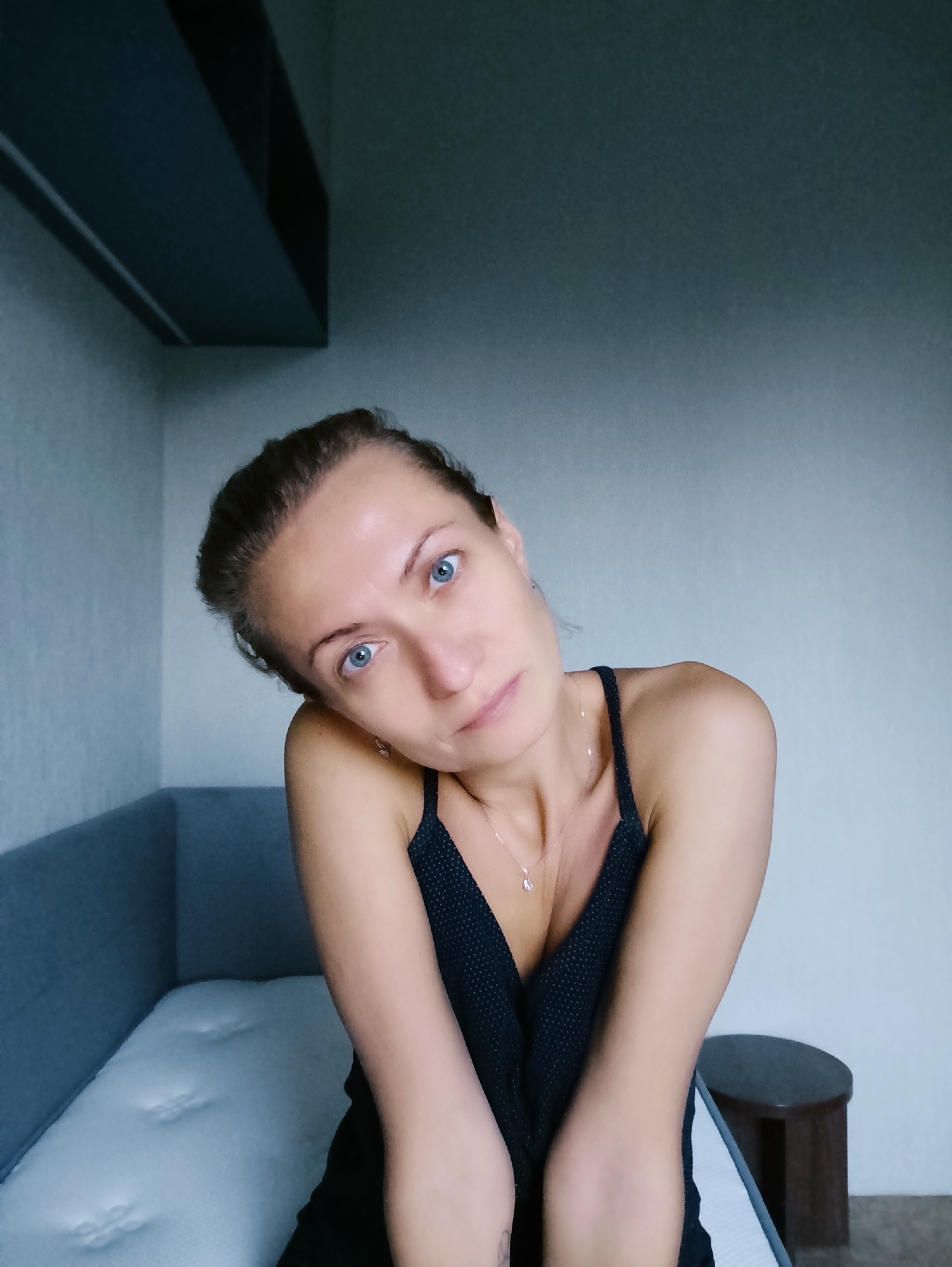In a Moscow apartment, a woman with blue eyes and her hair pulled back is captured in a selfie. She is wearing a black polka dot dress and a delicate necklace. Her head is tilted, and her arms are crossed in front of her. She is seated on the edge of a bed, with a gray upholstered headboard behind her, and a small, dark wooden stool to her right. The room appears minimalist, with plain walls and a dark, recessed ceiling fixture. The lighting is soft, creating a slightly diffused atmosphere.